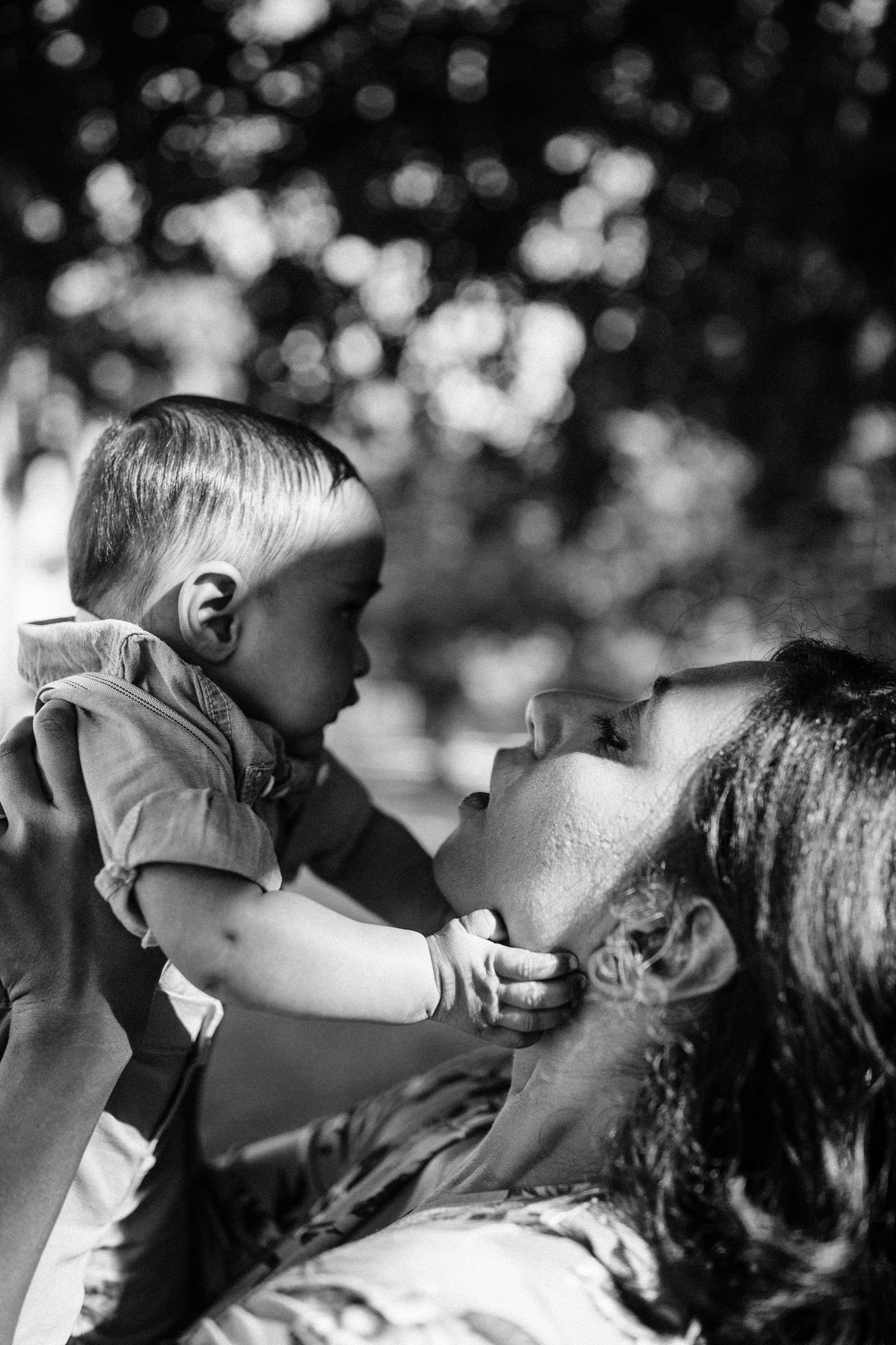 A toddler in profile gazes toward an adult while small hands grasp gently, dappled sunlight filtering through overhead foliage