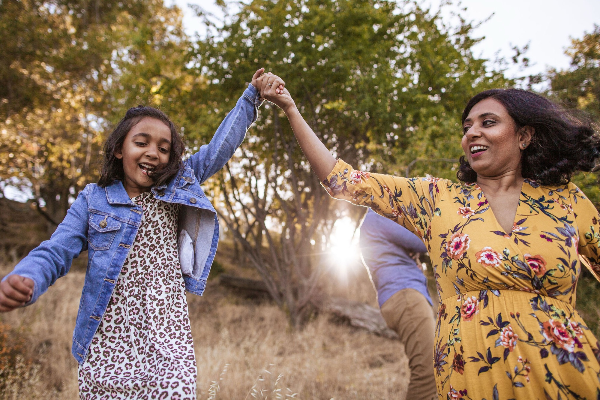 A woman and girl raise their clasped hands in joy along a tree-lined path, backlit by golden afternoon sunlight filtering through autumn foliage