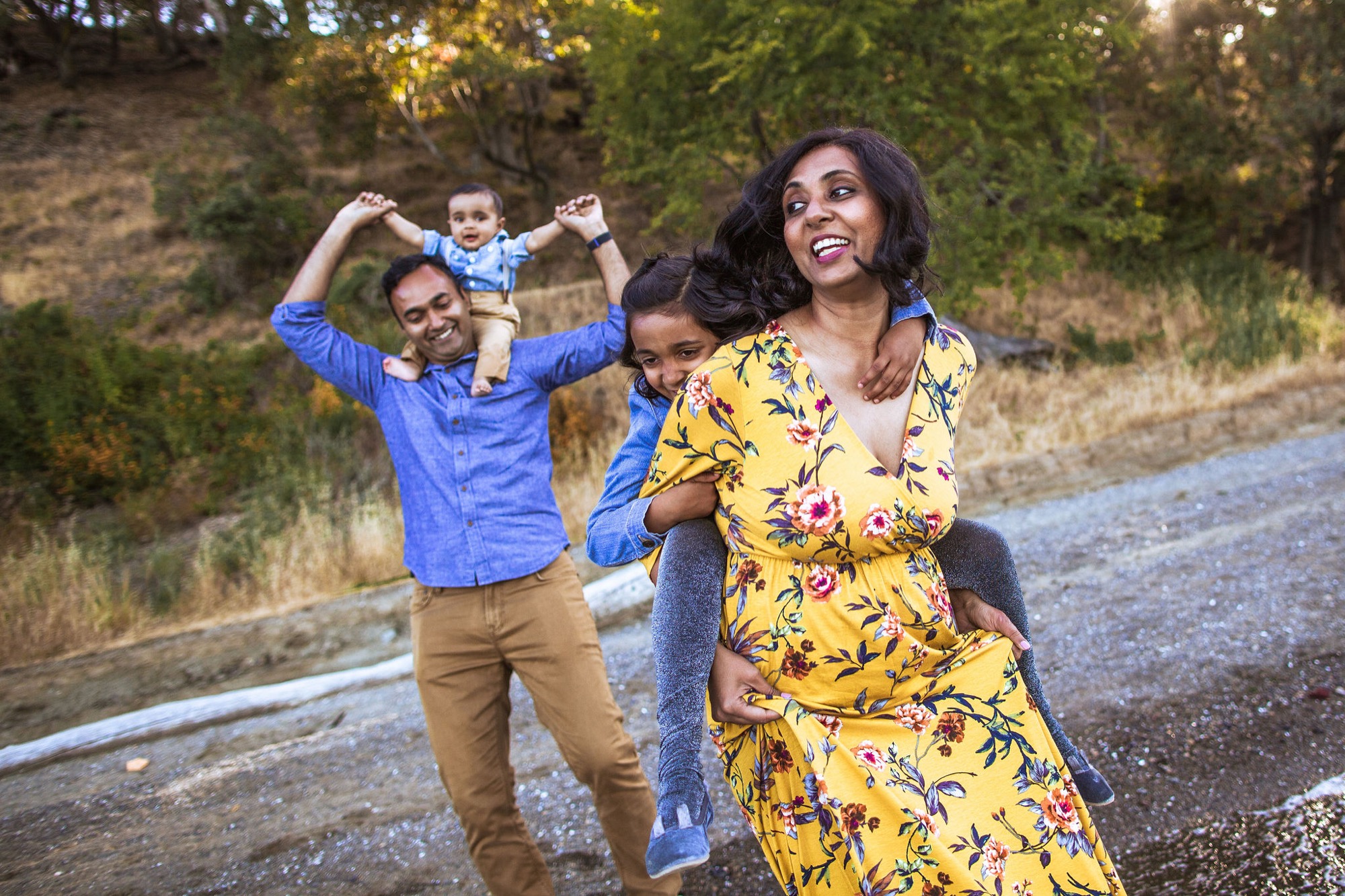 A joyful family laughs together on a gravel path, with the father lifting their toddler overhead while the mother and older daughter beam in the golden afternoon light