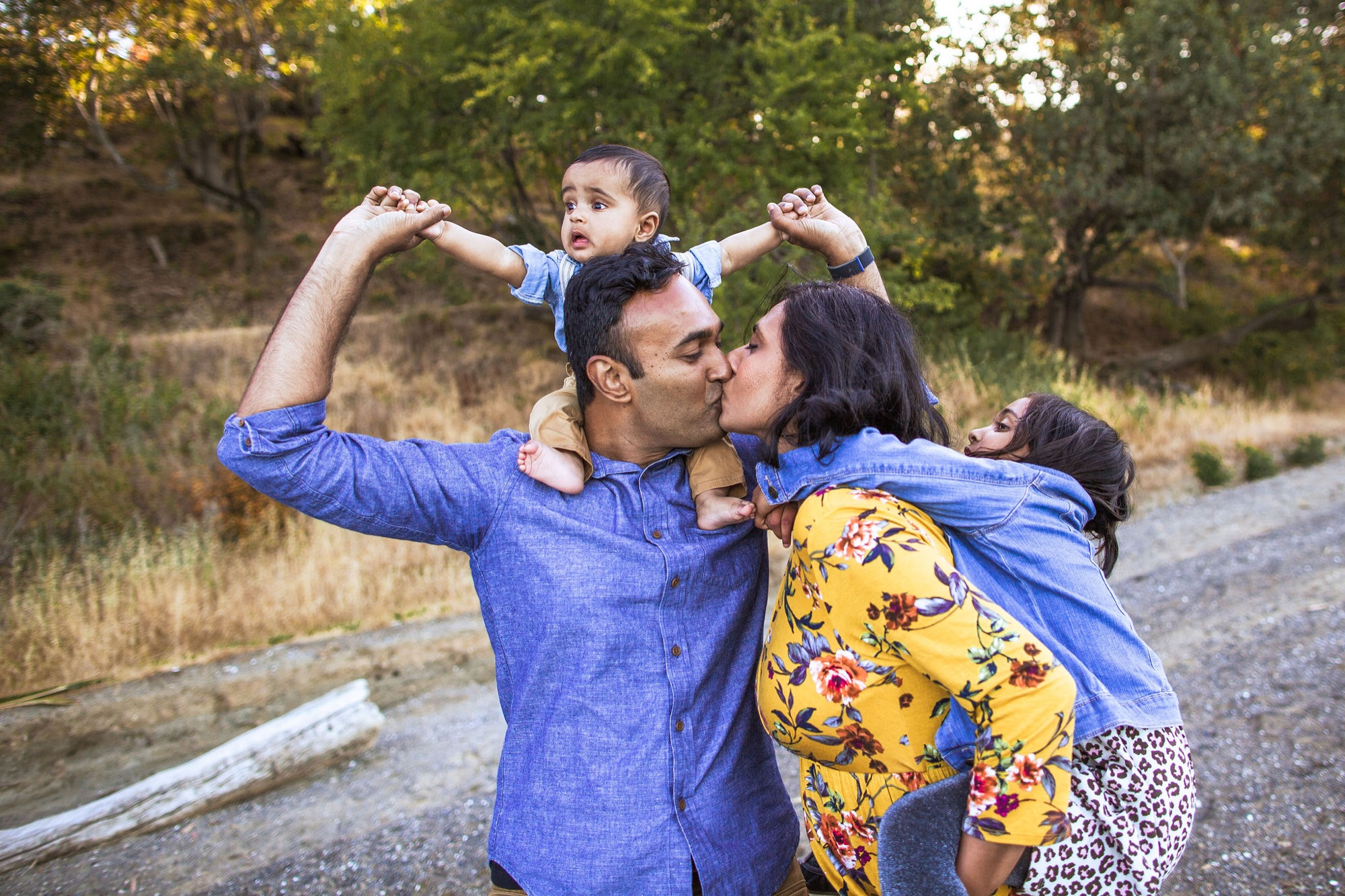 Father holds toddler overhead while parents kiss on a tree-lined forest path, capturing genuine joy and connection