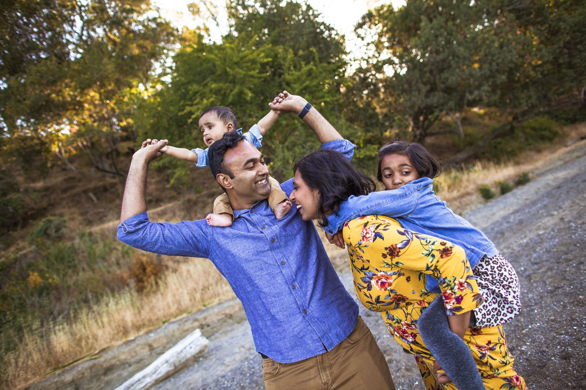 A father lifts his toddler overhead while his wife and older daughter lean in close, laughing together on a sun-dappled dirt road