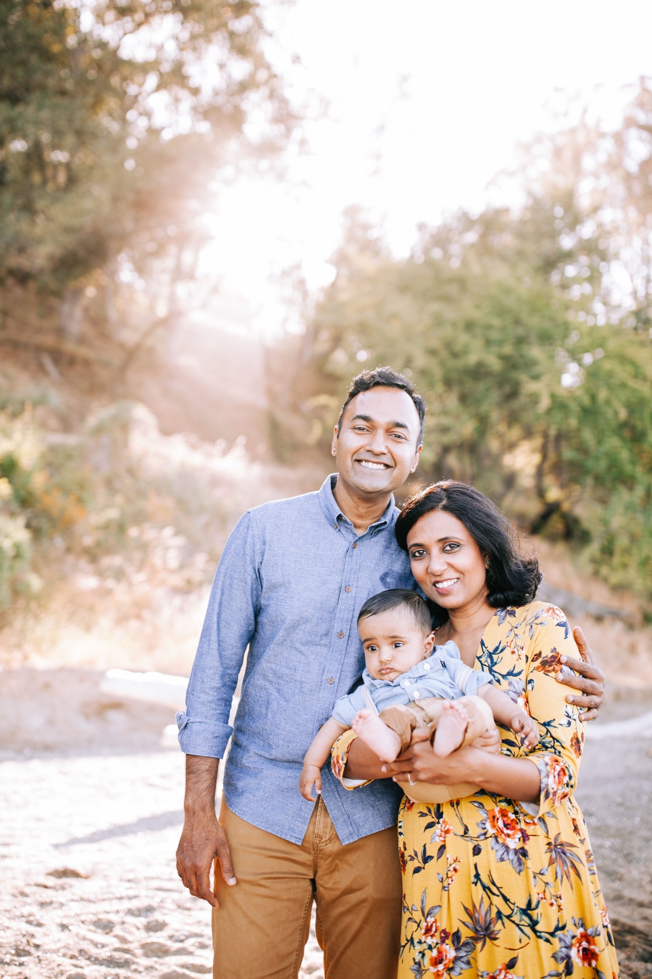 A family of three smiles warmly on a sunlit path lined with trees, the father in blue shirt and mother in yellow floral dress holding their infant son