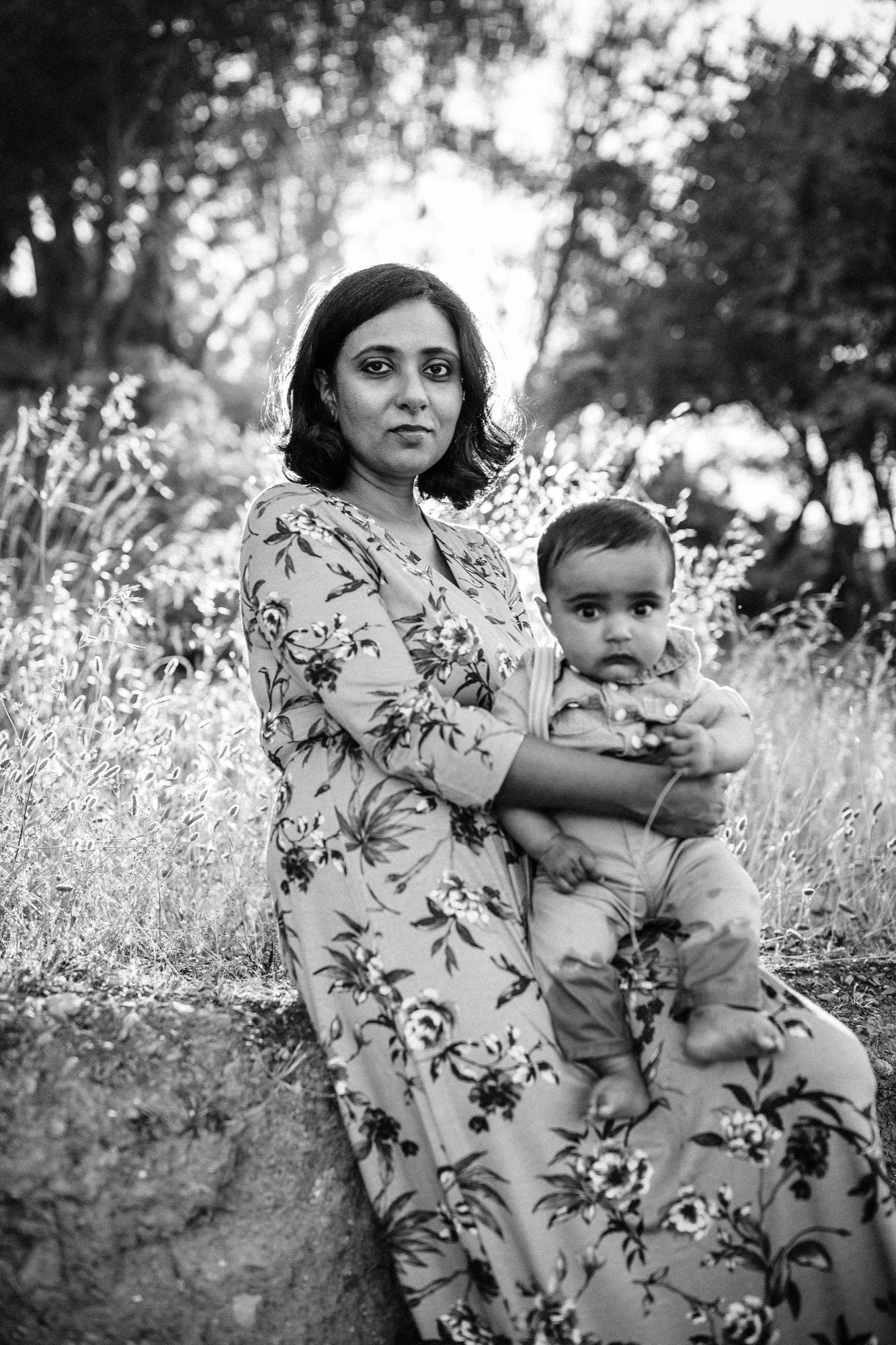 A woman in a floral dress holds her young child steady among wildflowers, both gazing directly at the camera with serene expressions