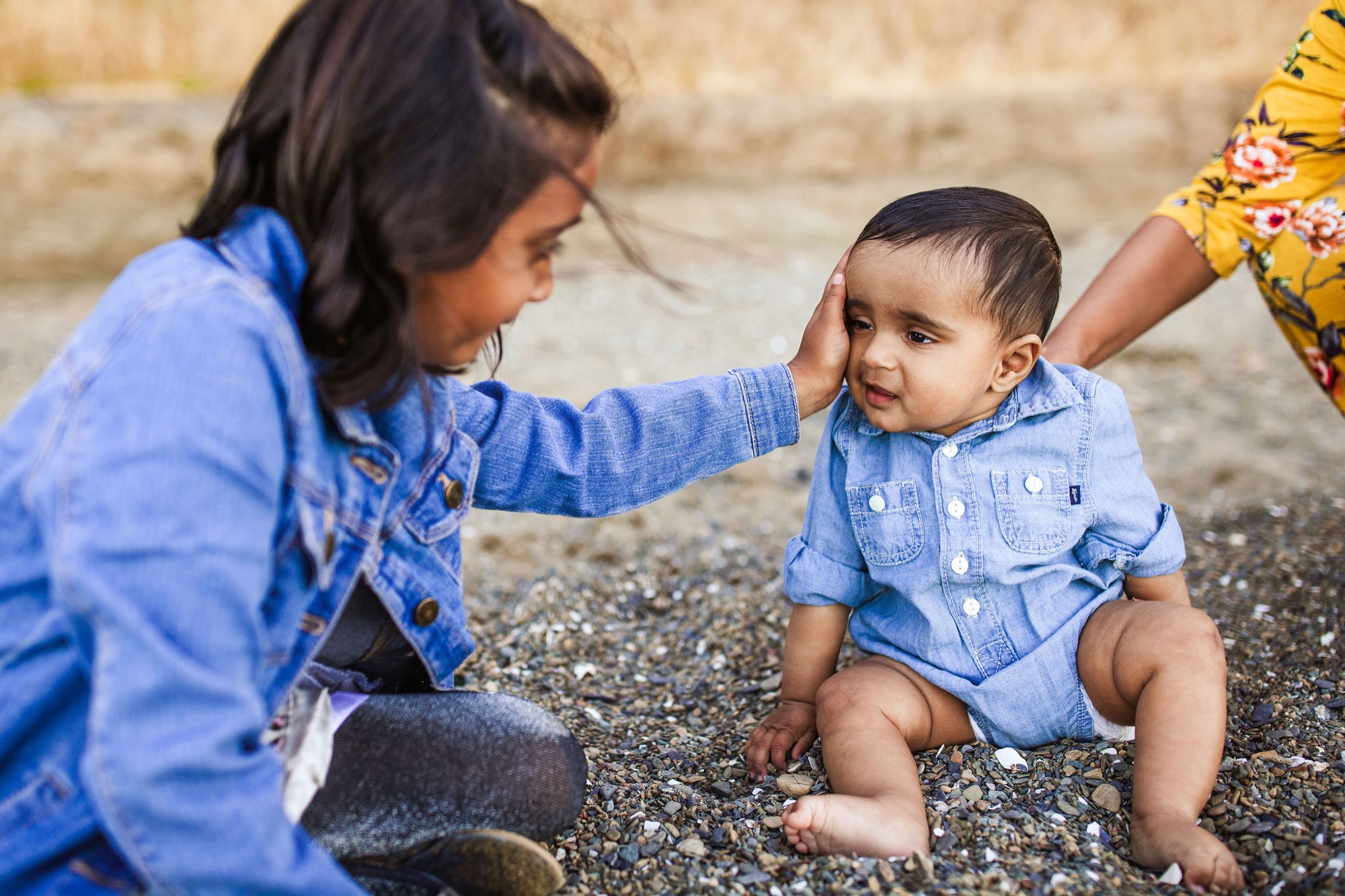 A young girl in denim gently touches a toddler's face as he sits on gravel, both wearing matching blue shirts