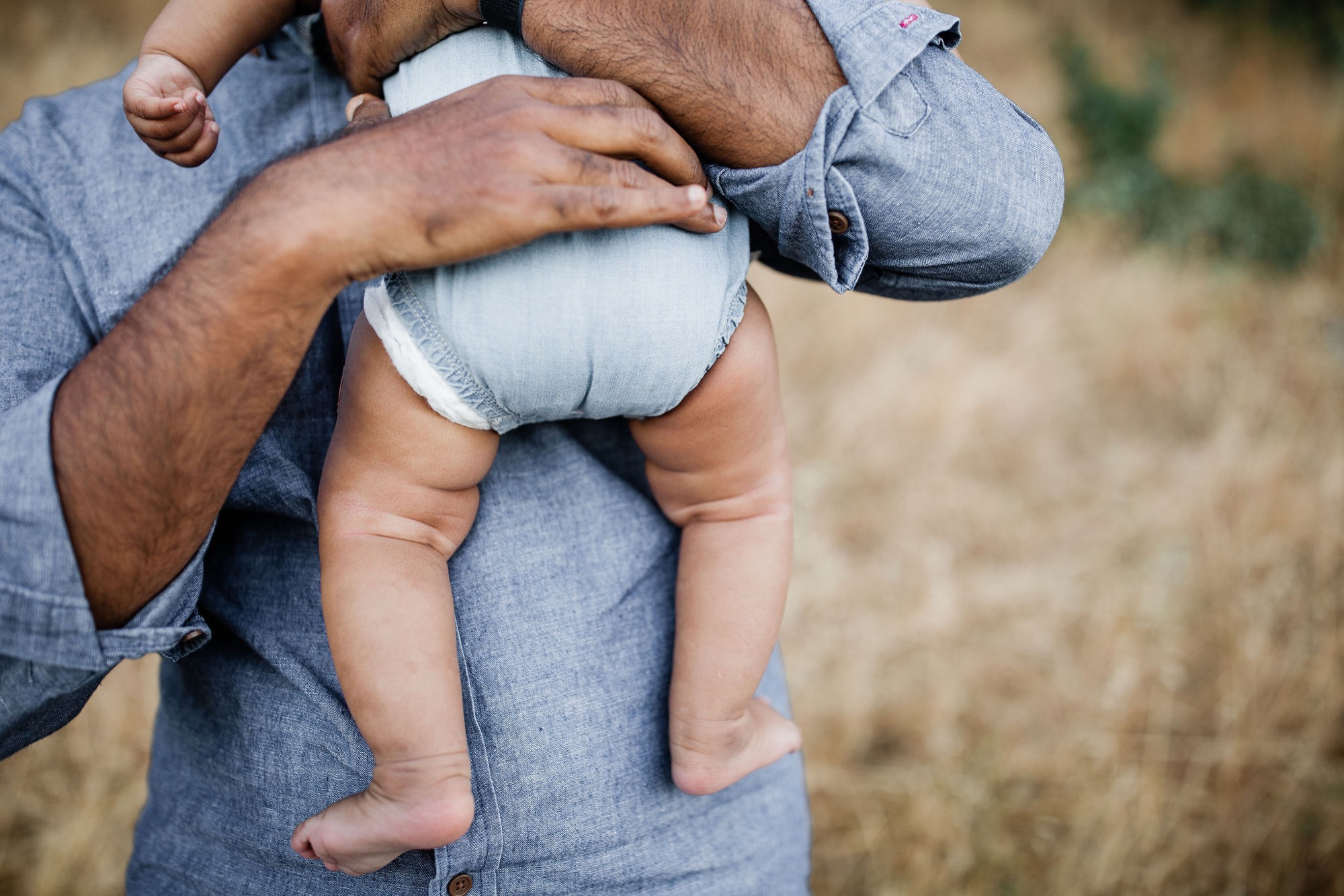 A father in denim cradles his infant son against his chest, the baby's small hand reaching upward in a tender moment of connection