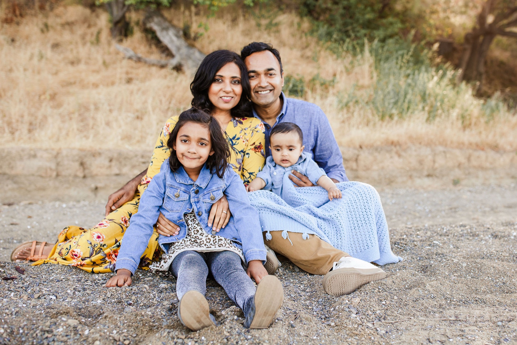A family of four sits closely together on sandy ground, dressed in coordinating blue and yellow tones, smiling warmly at the camera with natural hillside backdrop