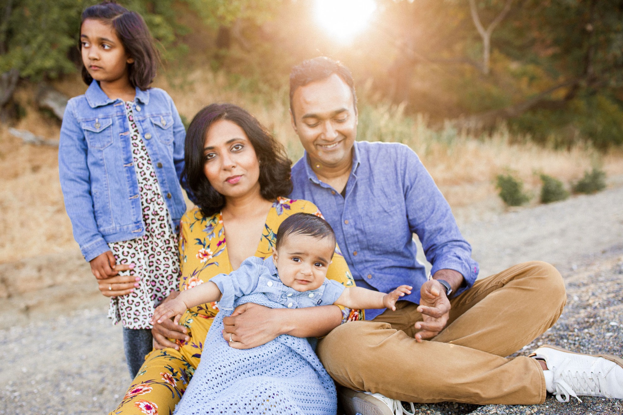 A family of four sits together on a dusty trail, bathed in golden sunlight, with the older daughter standing behind while parents embrace their toddler