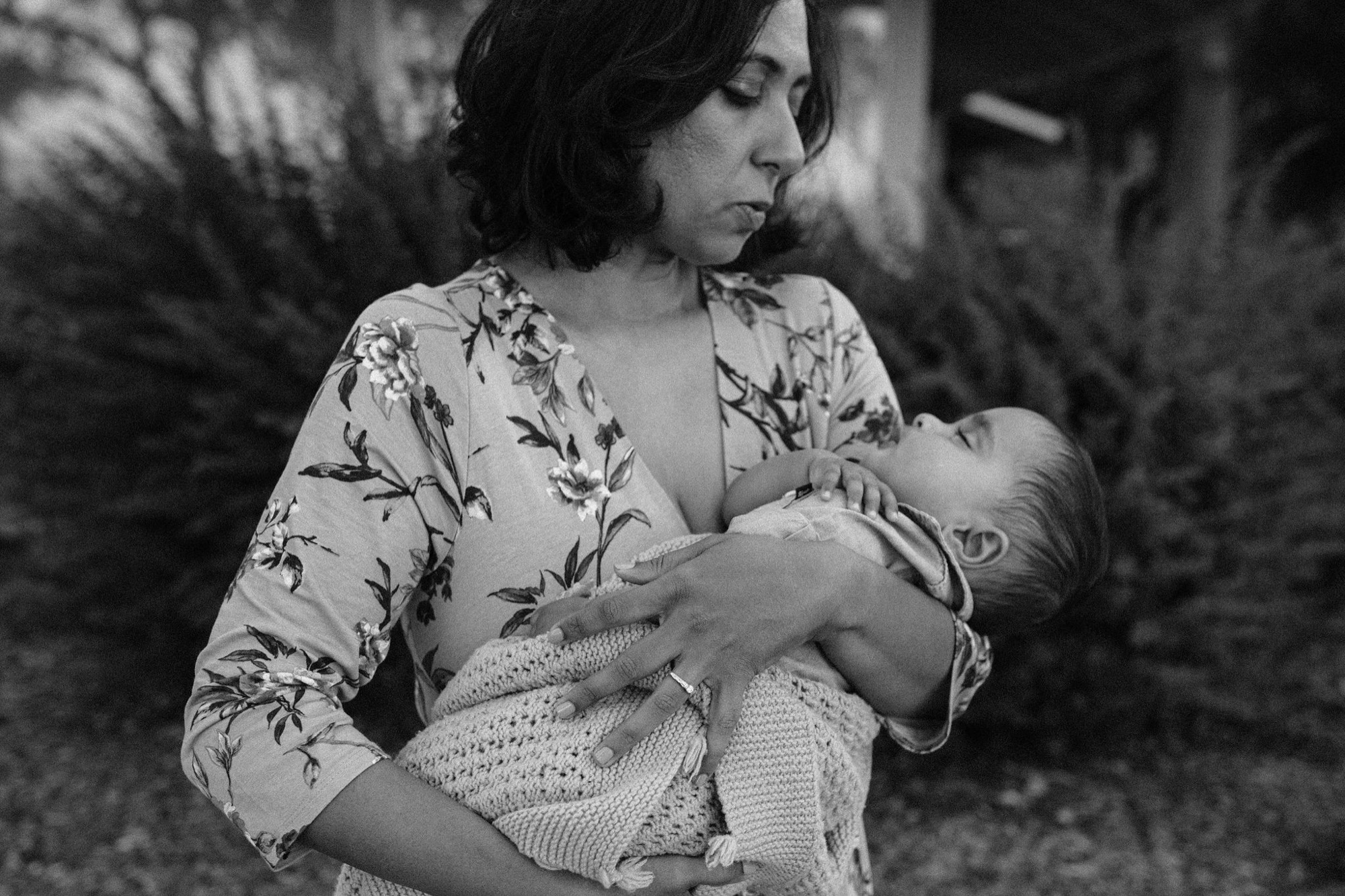 A mother in a floral dress holds her infant while gazing thoughtfully away, their profiles silhouetted against soft natural light in a wooded setting