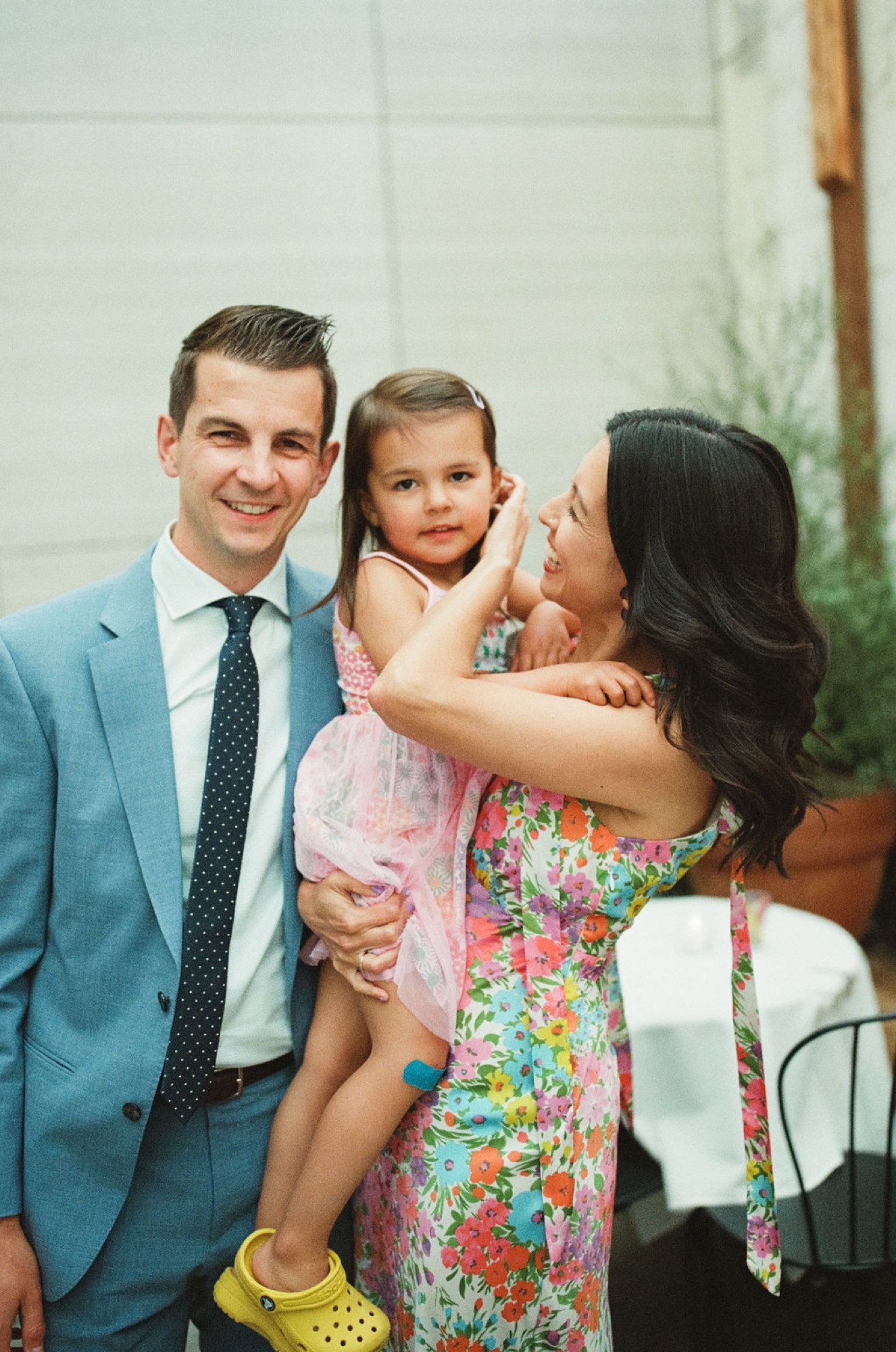 A family of three smiles warmly against a white brick wall, the mother holding their young daughter in a colorful floral dress