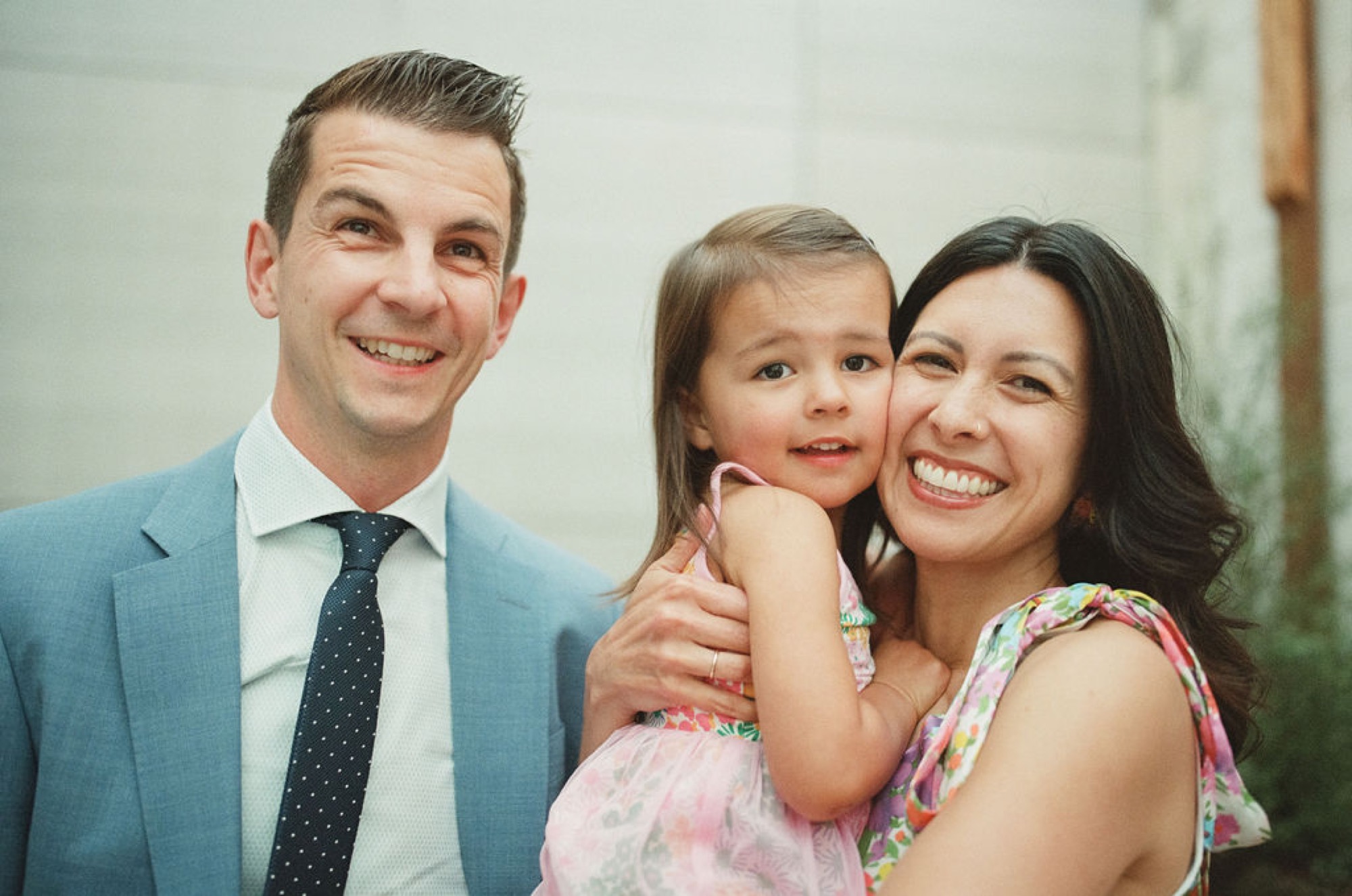 A smiling father in a light blue blazer stands beside his wife and young daughter in a pink floral dress, all beaming at the camera