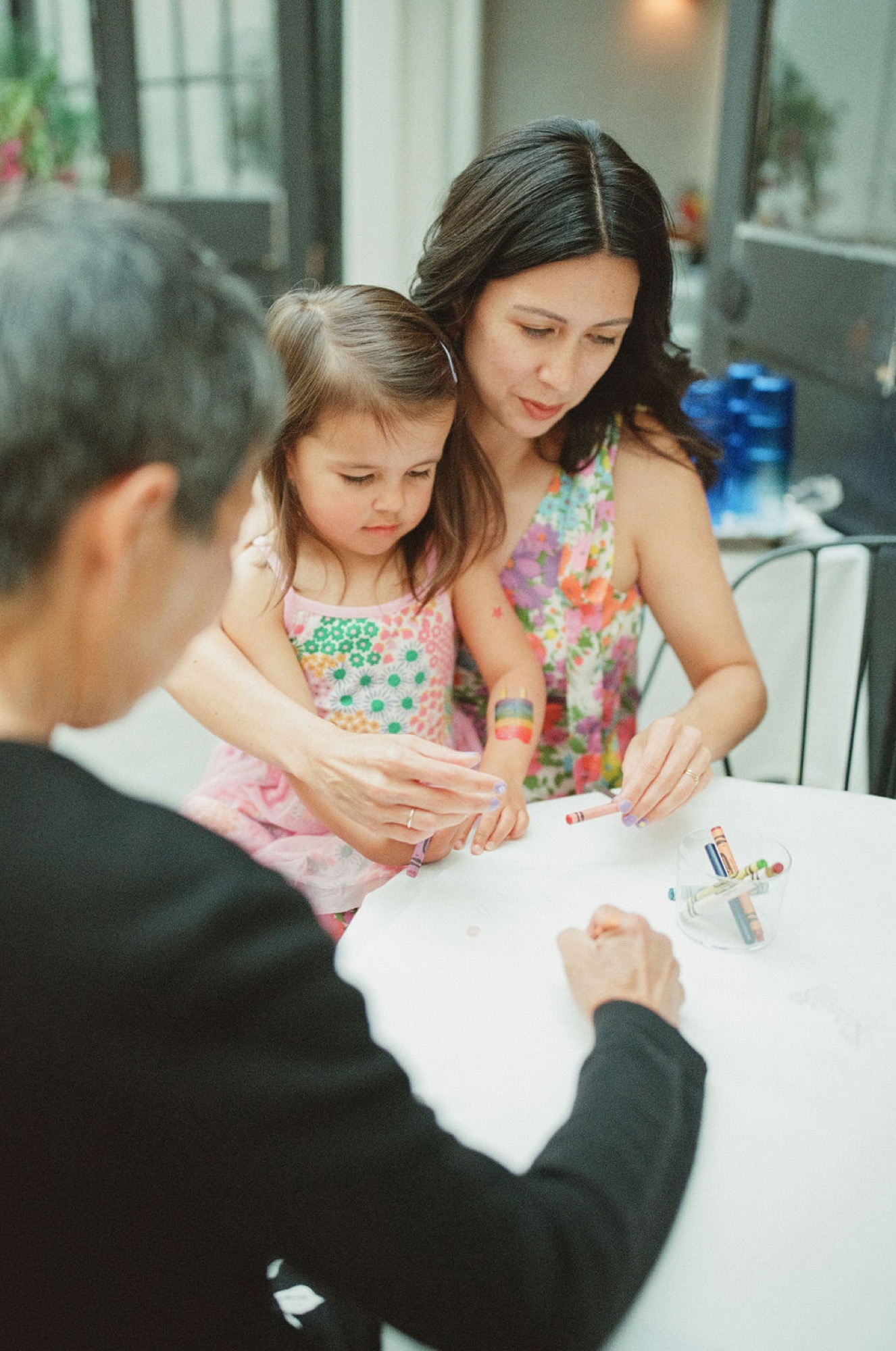 A woman and young girl lean over a white table, concentrating intently as they color together with crayons indoors