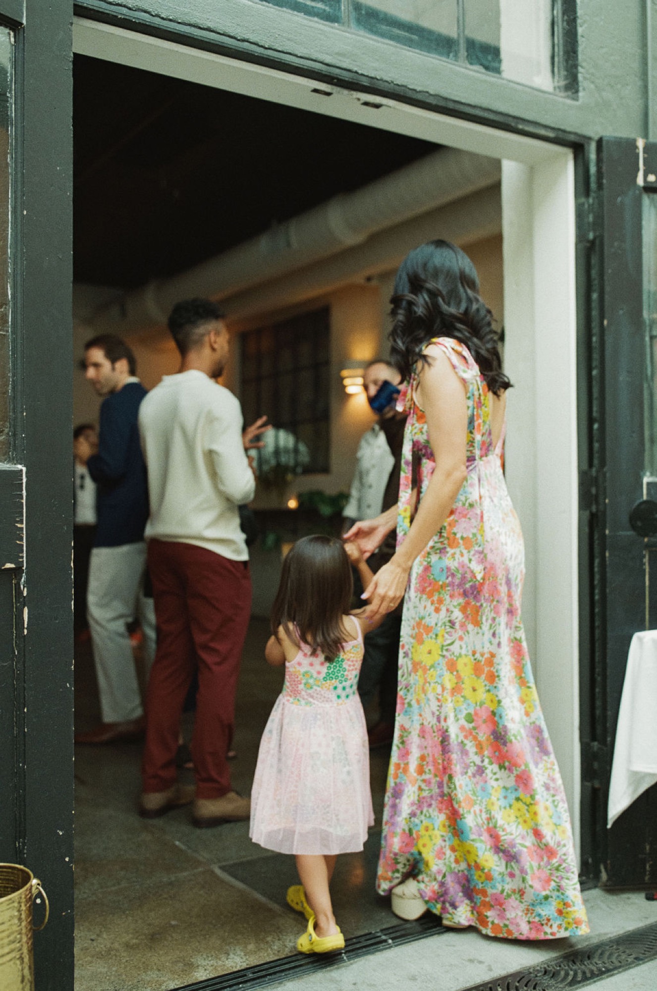 A woman in a vibrant floral dress guides her young daughter by the hand into a warmly lit home, while guests gather inside
