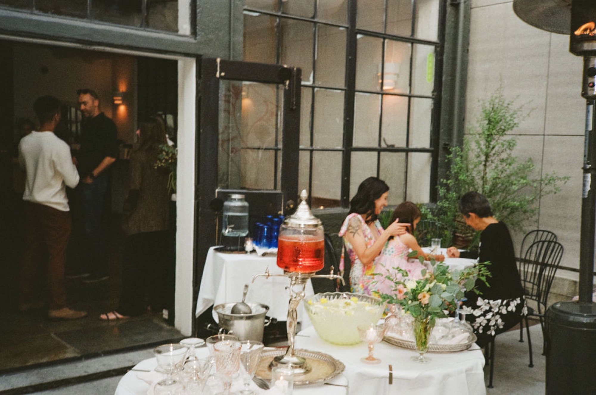 A woman in a floral dress embraces a child while seated at an outdoor dinner table, with guests mingling near the entrance behind her