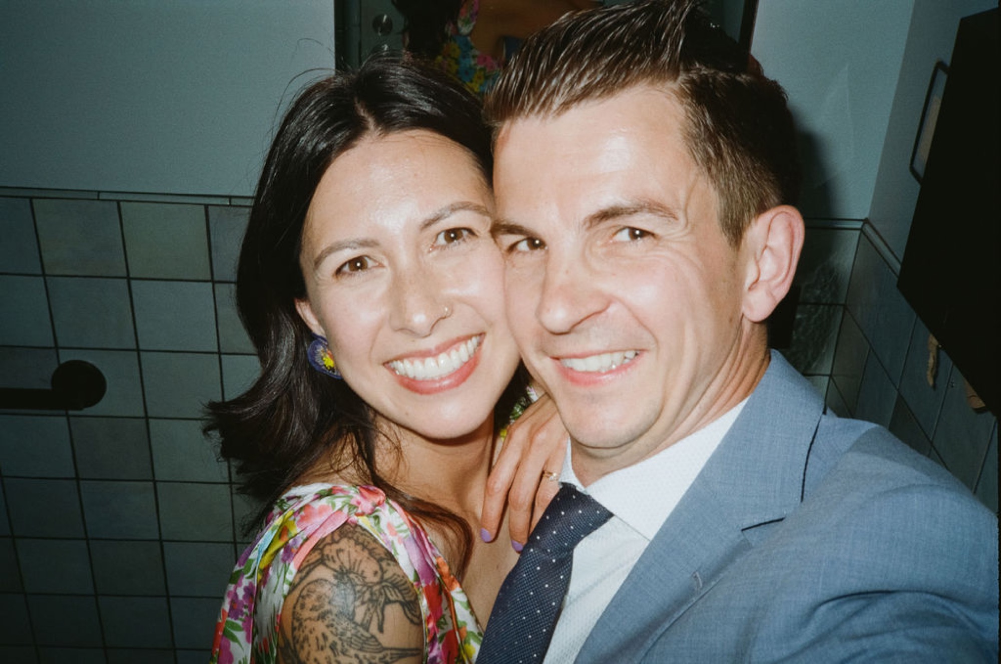 A couple smiles warmly at the camera in an indoor setting, their faces close together in an intimate moment of genuine joy and connection