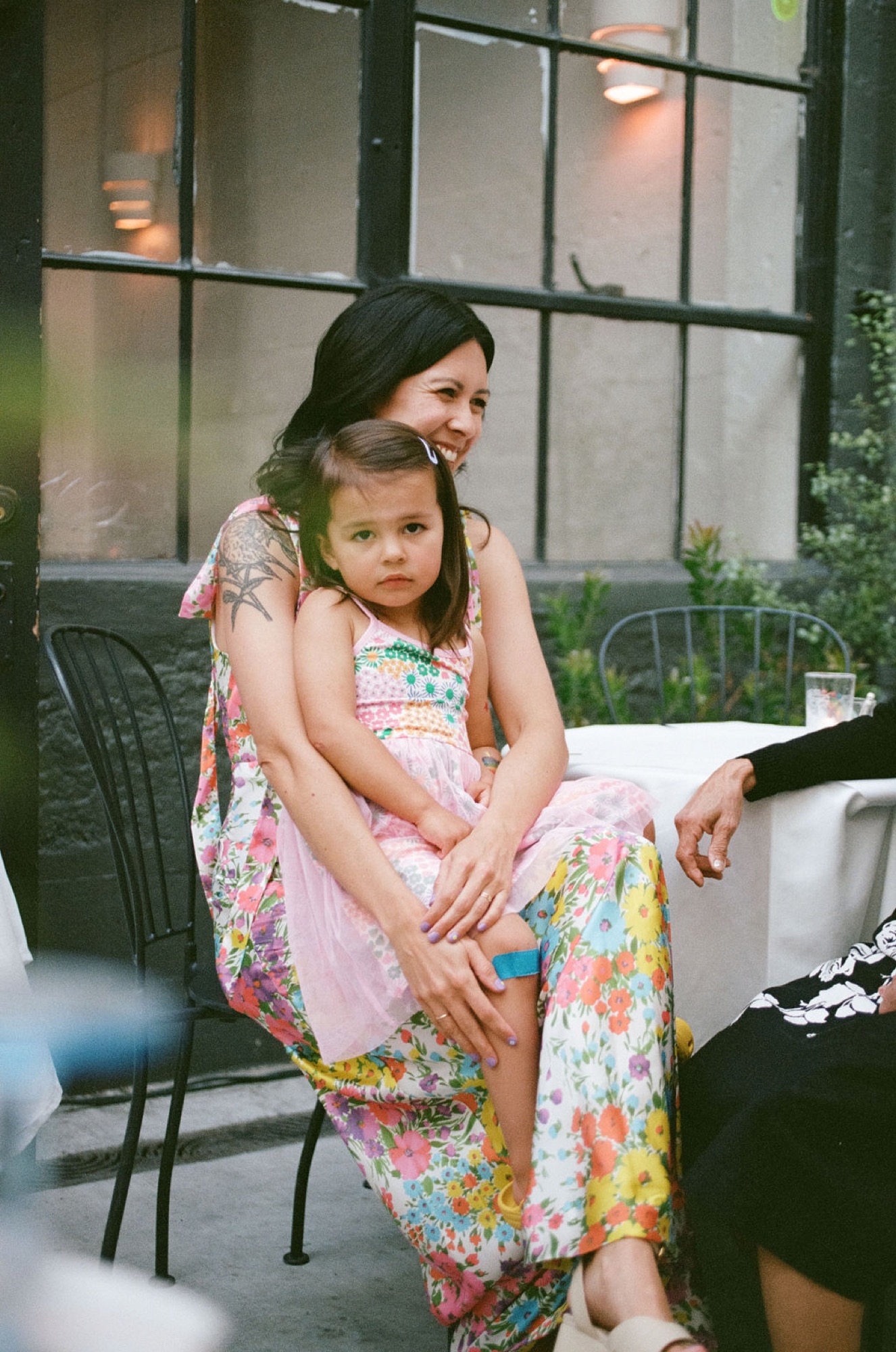 A woman embraces her young daughter from behind on a patio, both wearing matching floral dresses, with soft afternoon light illuminating their warm connection