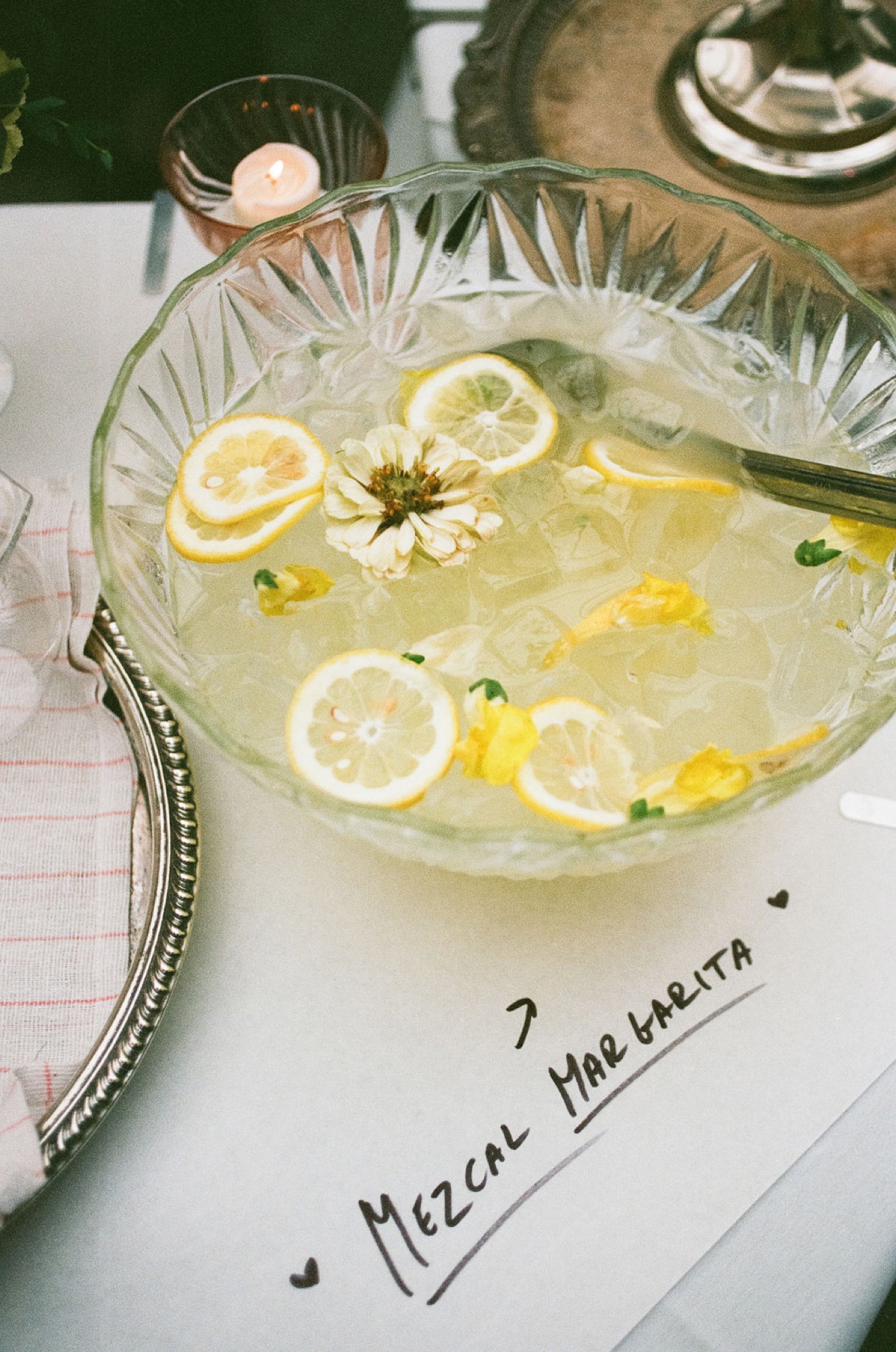 A pale green cocktail garnished with lemon slices and a white daisy flower sits in a decorative glass bowl labeled Mezcal Margarita
