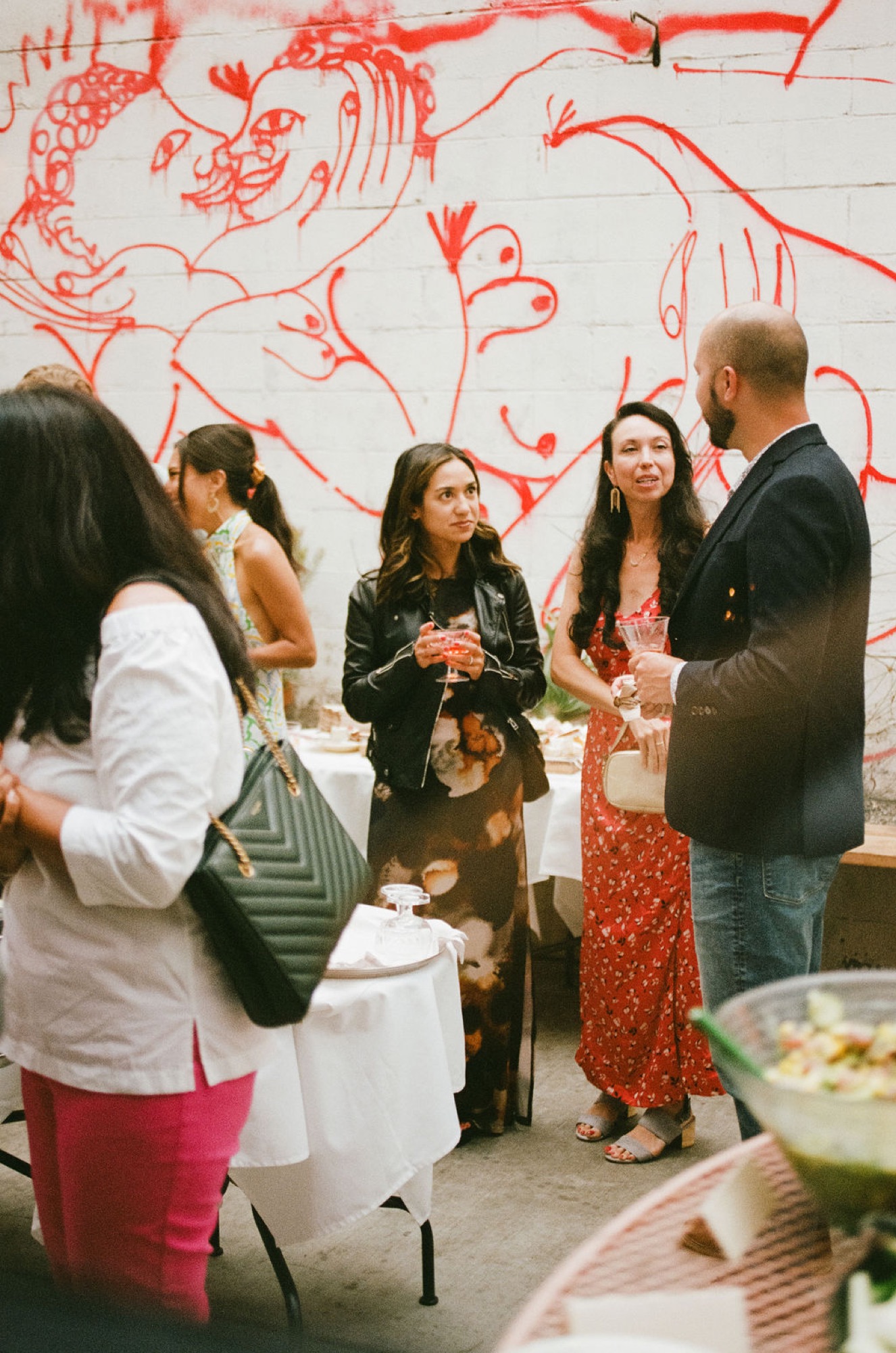 A group of adults socializes in front of a vibrant red mural, holding drinks and engaged in conversation at an indoor gallery or event space