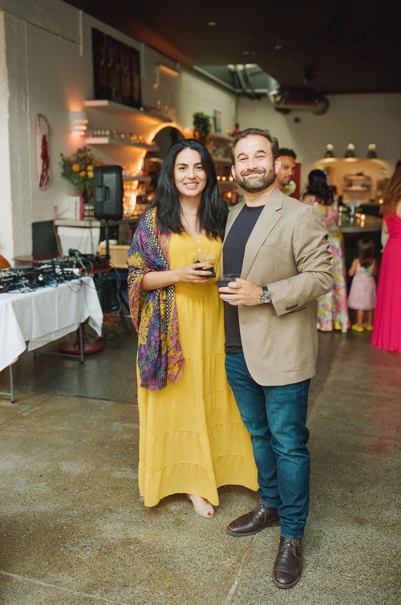 A smiling couple holds drinks at an indoor event, warmly illuminated by warm ambient lighting and decorative elements behind them