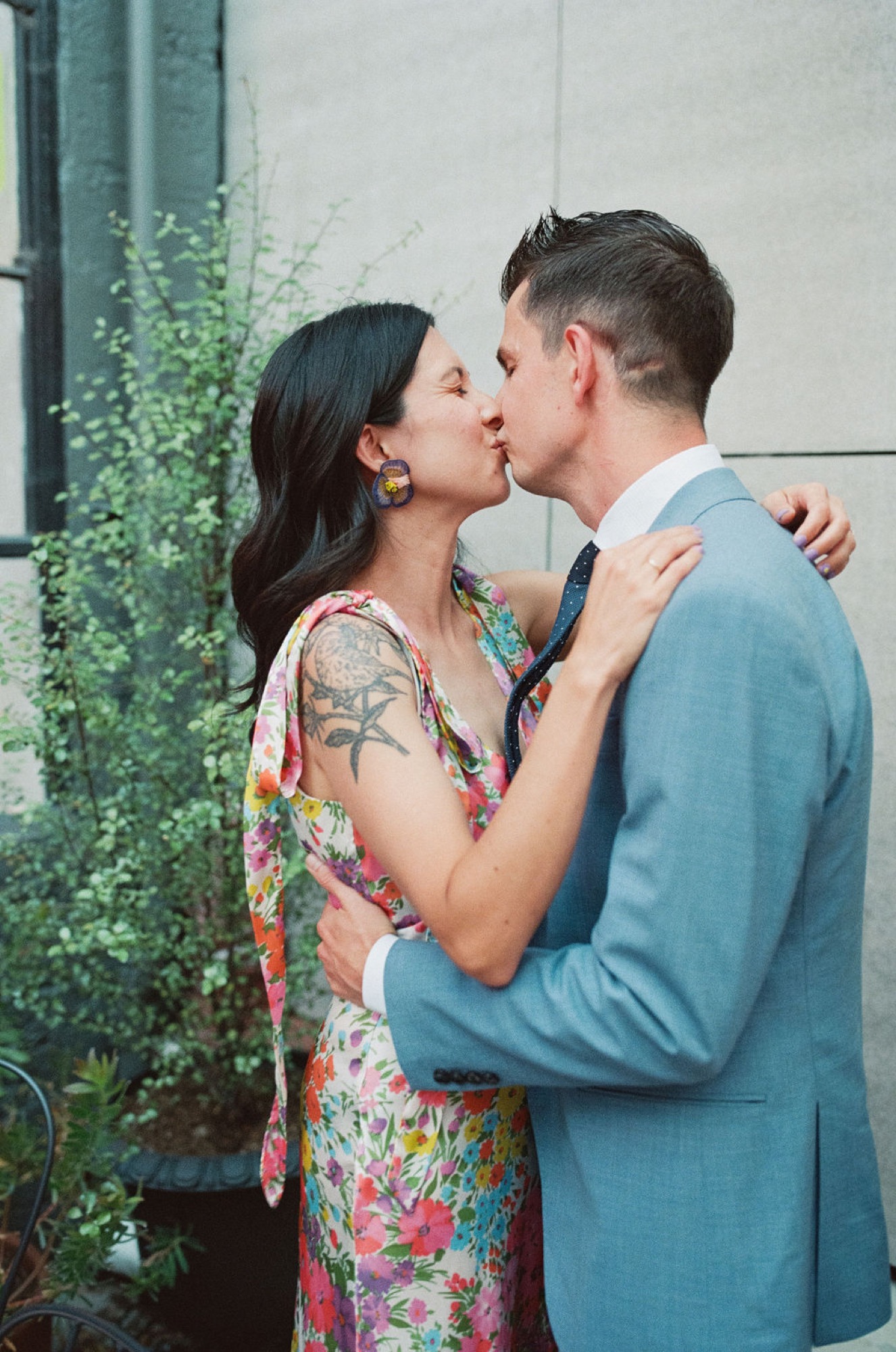A couple kisses tenderly against a white wall with climbing ivy, she in a colorful floral dress and statement earrings, he in a blue blazer