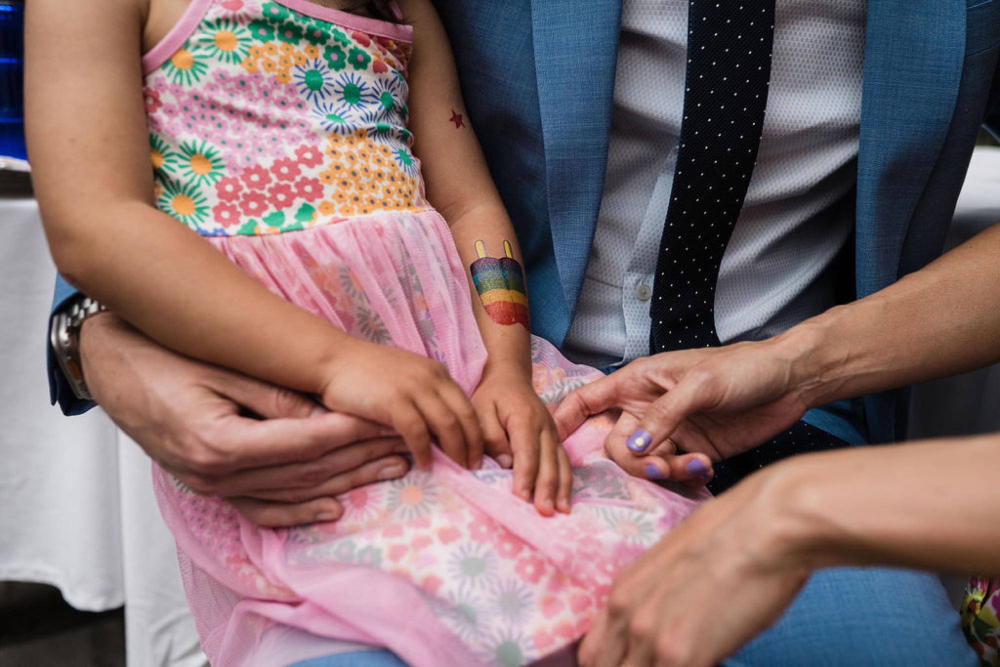 Multiple hands gently rest on a child's pink patterned dress, showing tender family connection and togetherness