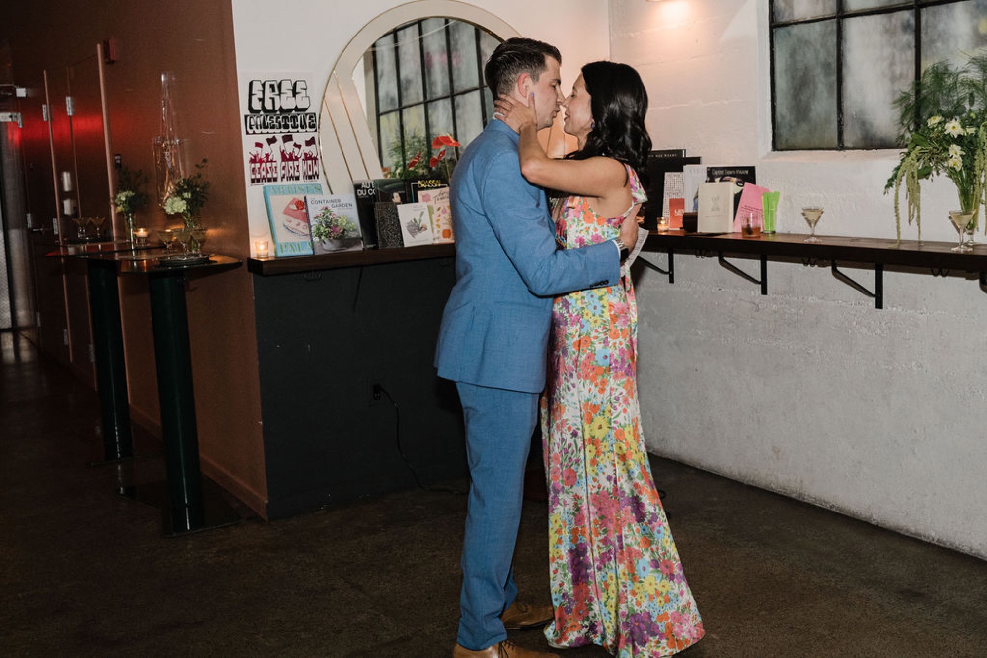 A couple shares an intimate kiss at an indoor venue, the man in a blue suit and woman in a vibrant floral dress, with decorated shelves and warm lighting behind them