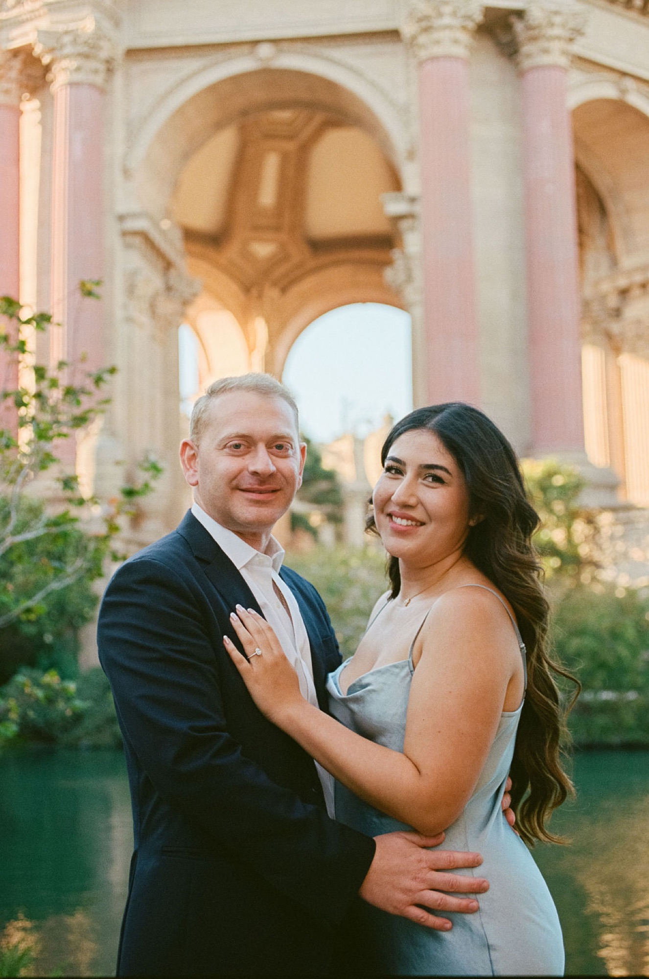 A couple smiles warmly at the camera beneath the Palace of Fine Arts' iconic arched colonnade, with soft natural light illuminating their elegant attire