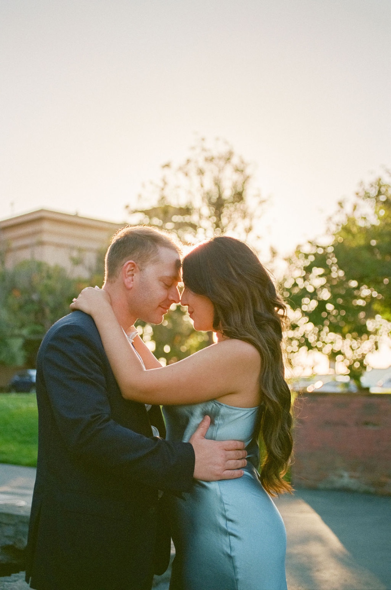 A couple shares an intimate moment at golden hour, foreheads nearly touching, with the Palace of Fine Arts and green grounds softly blurred behind them