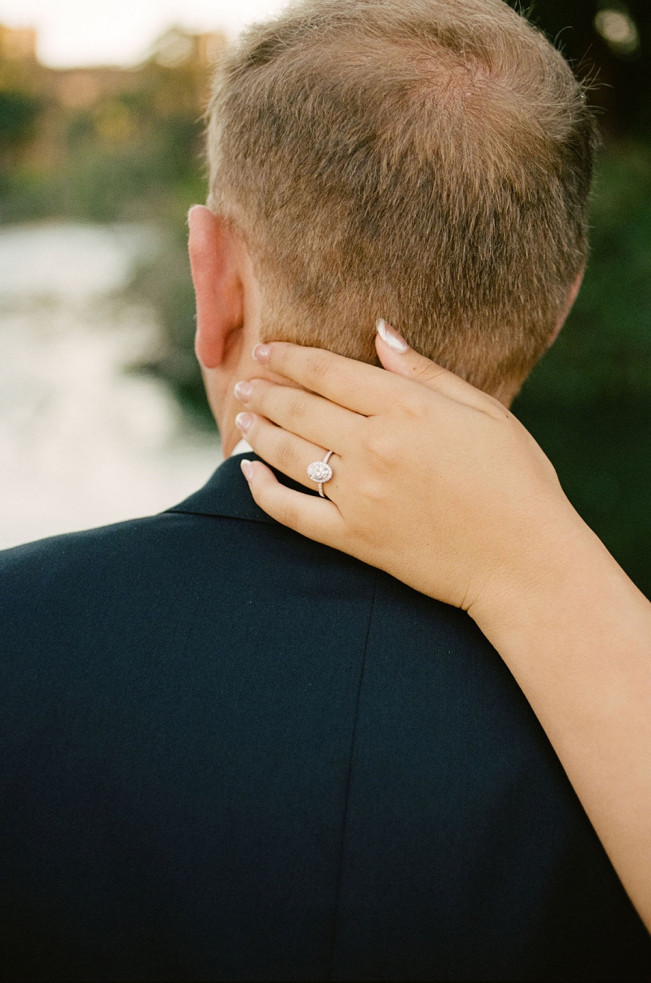 A woman's hand rests affectionately on a man's shoulder, her diamond ring catching warm golden light near water