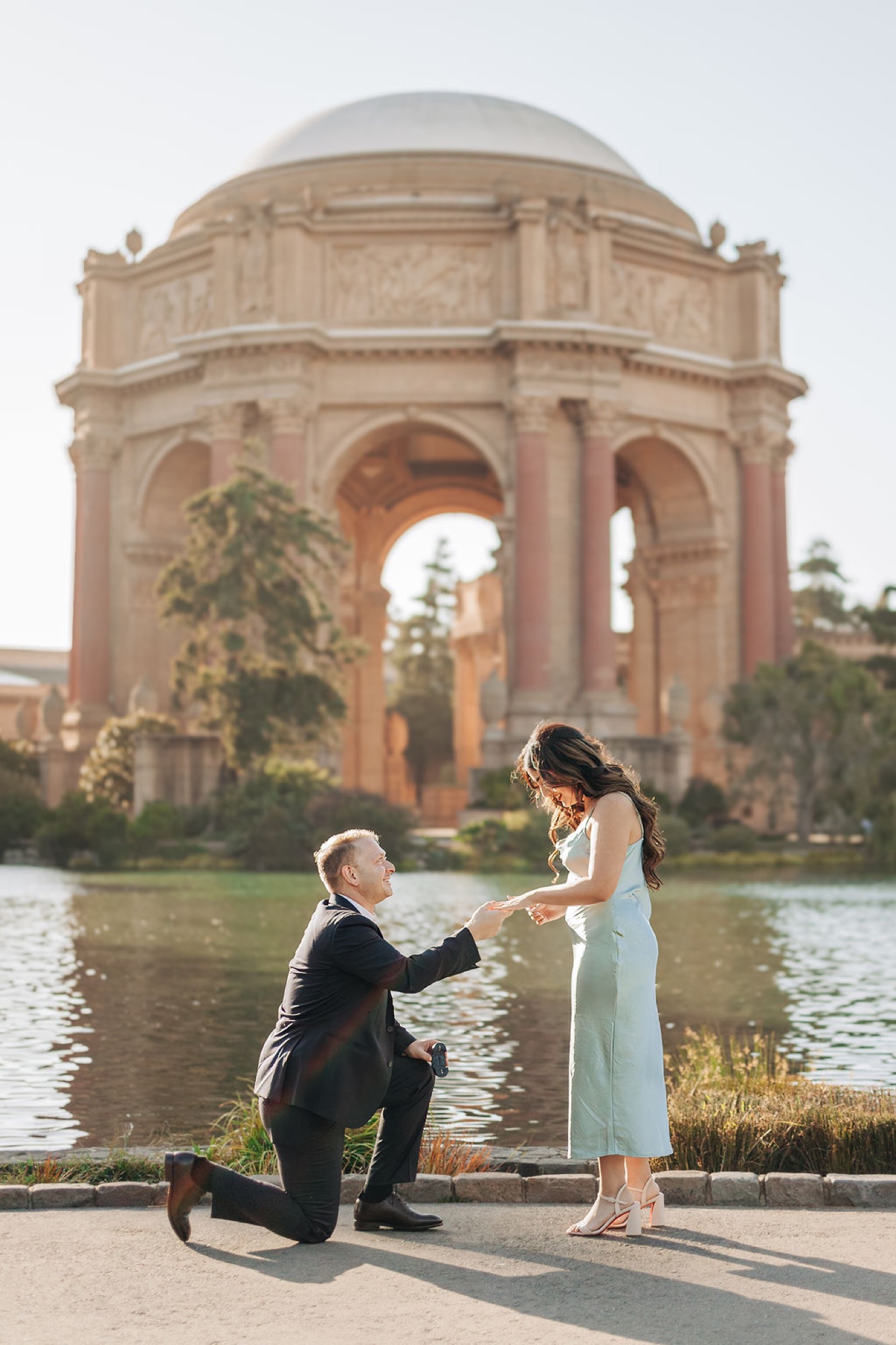 Man kneels on one knee proposing to a woman in a light dress beside the Palace of Fine Arts lagoon, the iconic dome rising behind them at golden hour