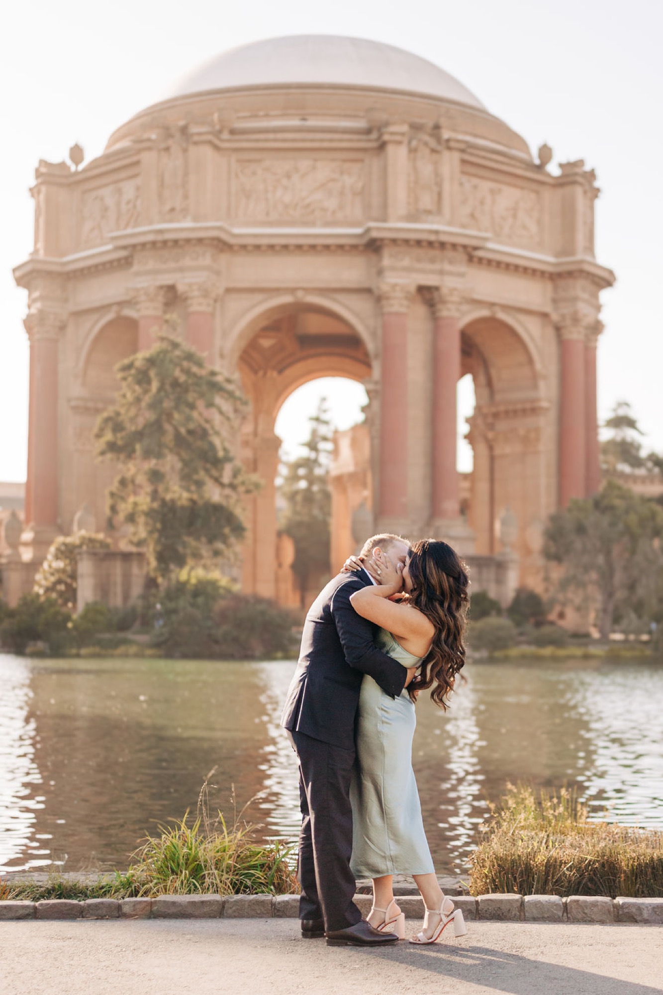A couple embraces and kisses in front of the Palace of Fine Arts, with the iconic dome and arches reflected in the still water below