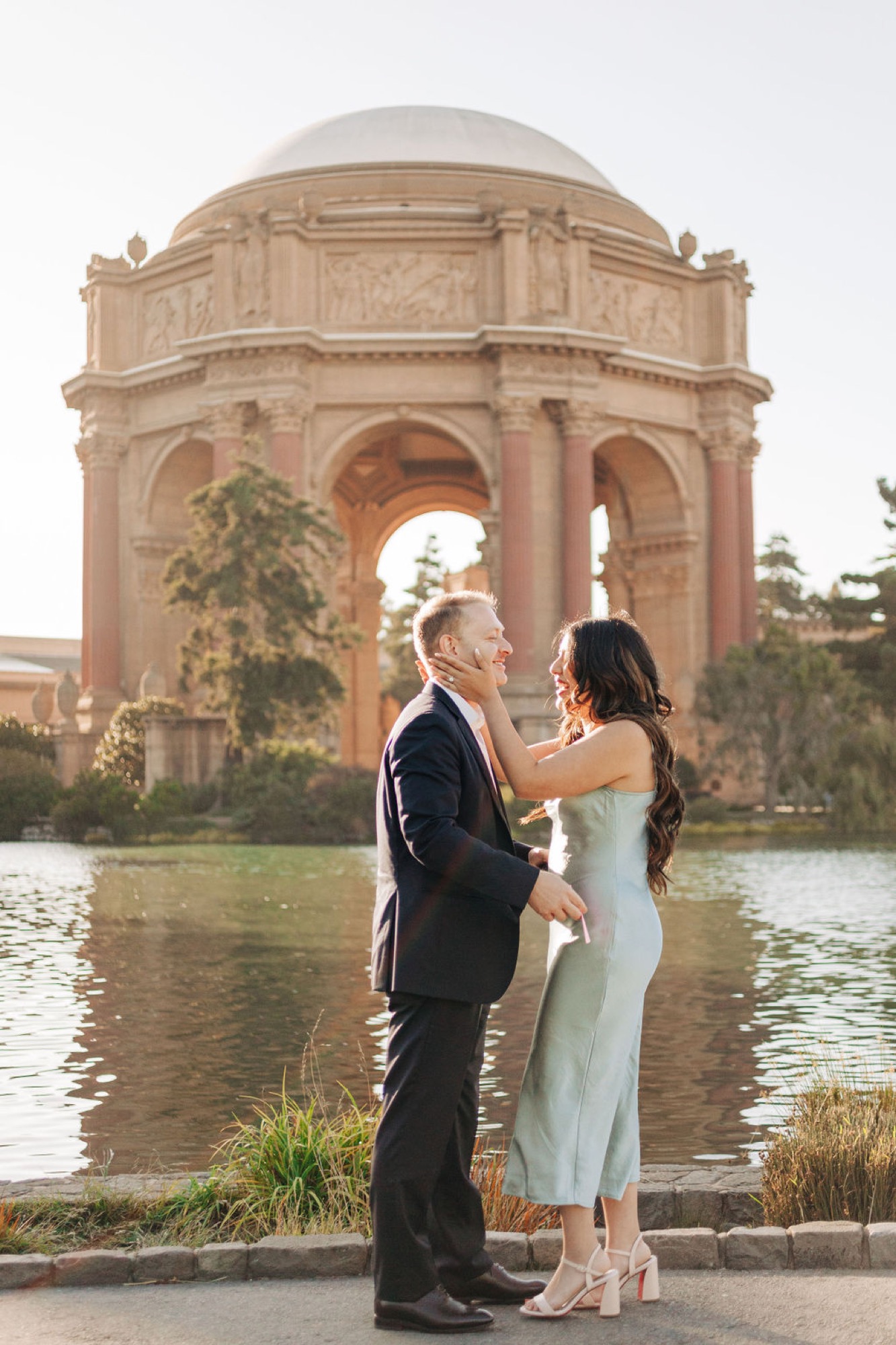 Couple in formal attire stands facing each other by the Palace of Fine Arts lagoon, soft afternoon light illuminating the iconic dome behind them