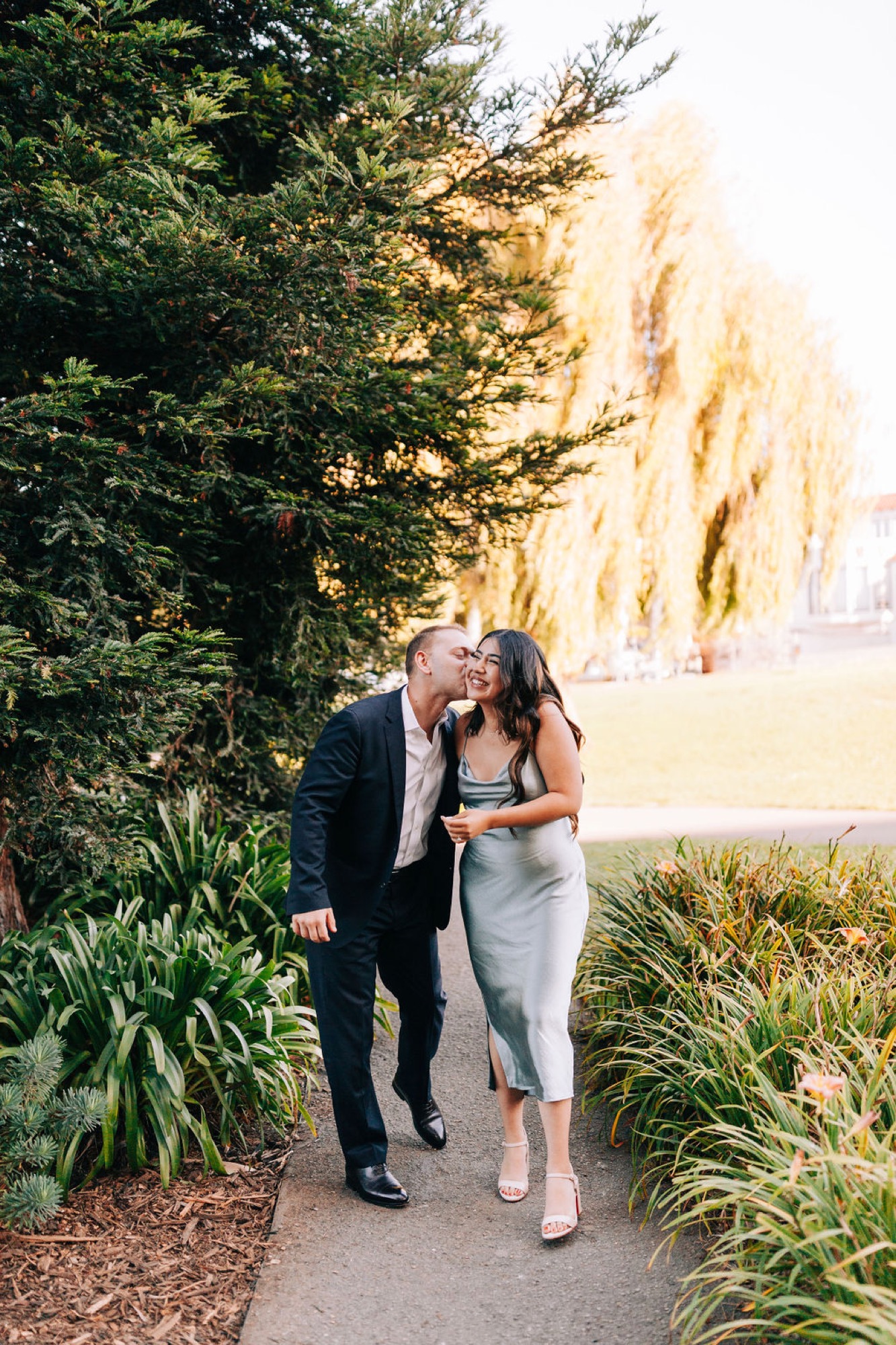 A couple walks arm-in-arm along a tree-lined path, with the man kissing the woman's forehead as golden afternoon light filters through the canopy