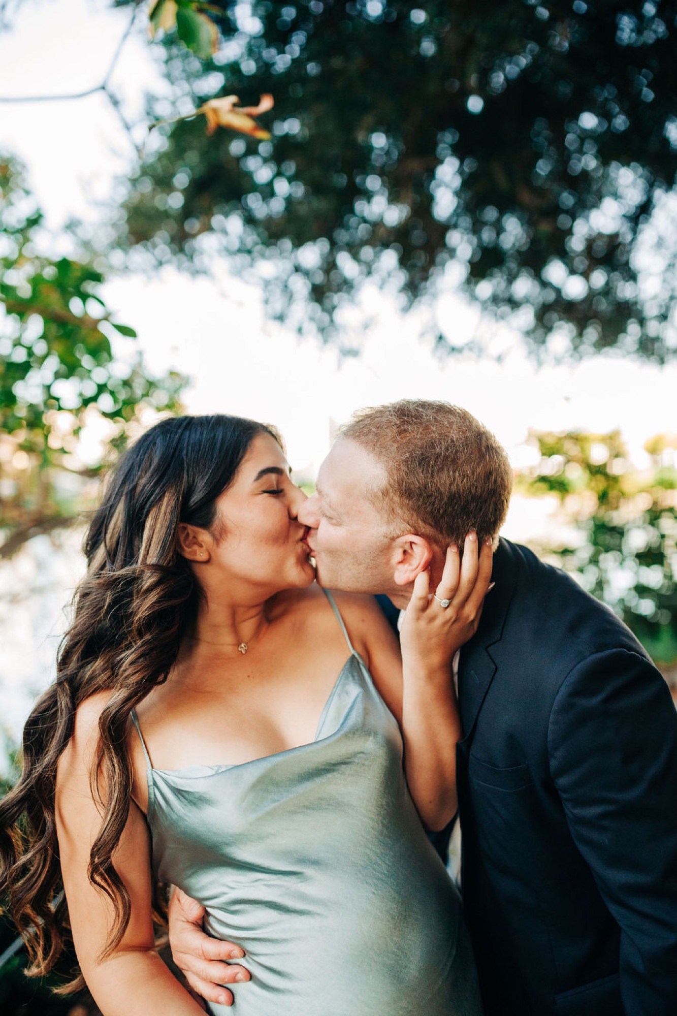 A couple shares an intimate kiss beneath dappled tree canopy with soft natural light filtering through leafy branches overhead