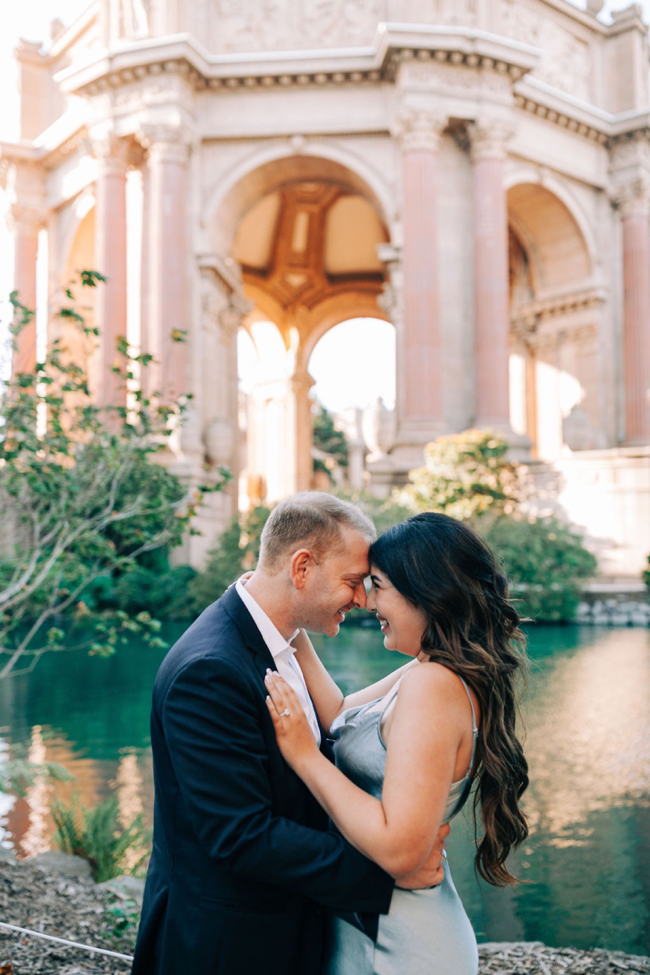 A couple shares an intimate moment at the Palace of Fine Arts, with golden arches and turquoise water framing their embrace in soft afternoon light