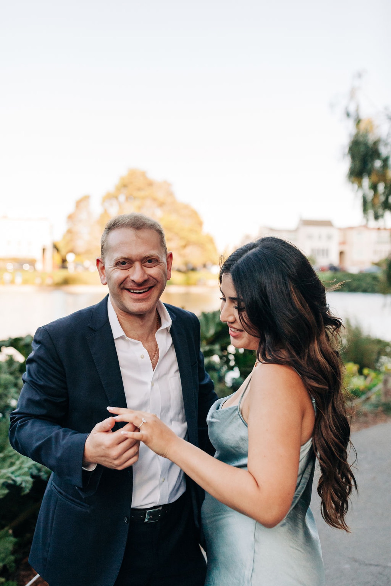 A man in a navy blazer and woman in a light blue dress laugh together at sunset, the Palace of Fine Arts glowing softly behind them
