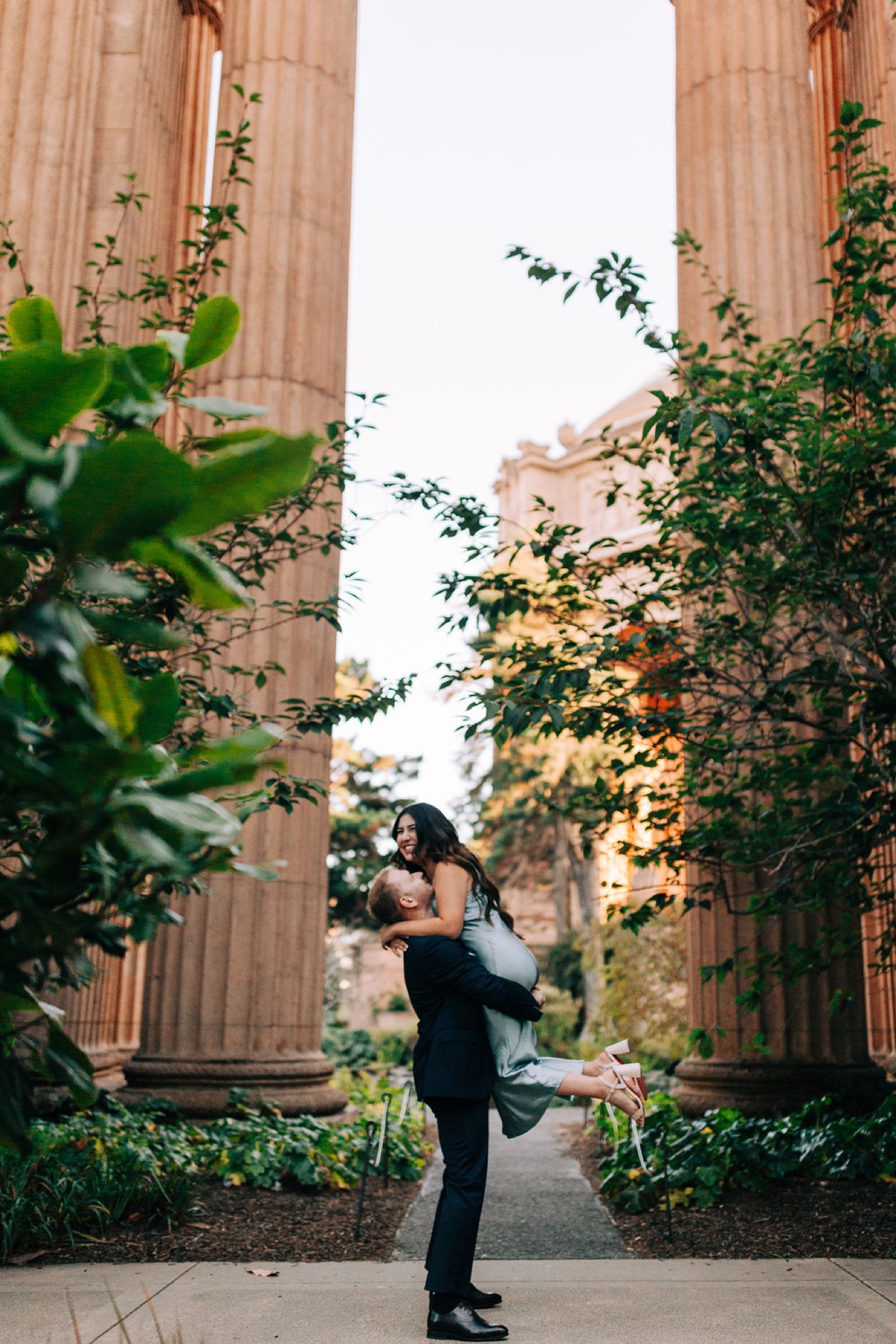 A woman in black clothing kicks up her leg playfully between two towering fluted columns, ivy climbing the classical architecture behind her