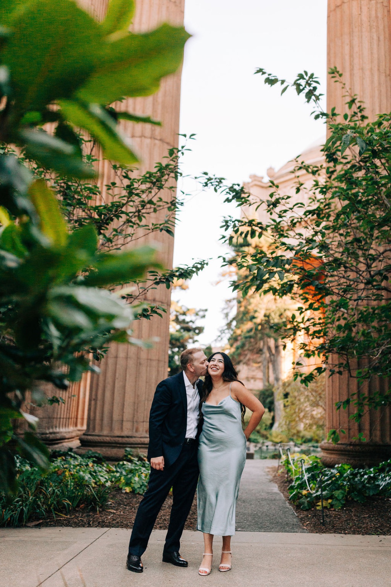 A couple in formal attire walks through a verdant urban corridor, ivy-draped brick buildings framing their intimate moment as soft golden light filters through the foliage
