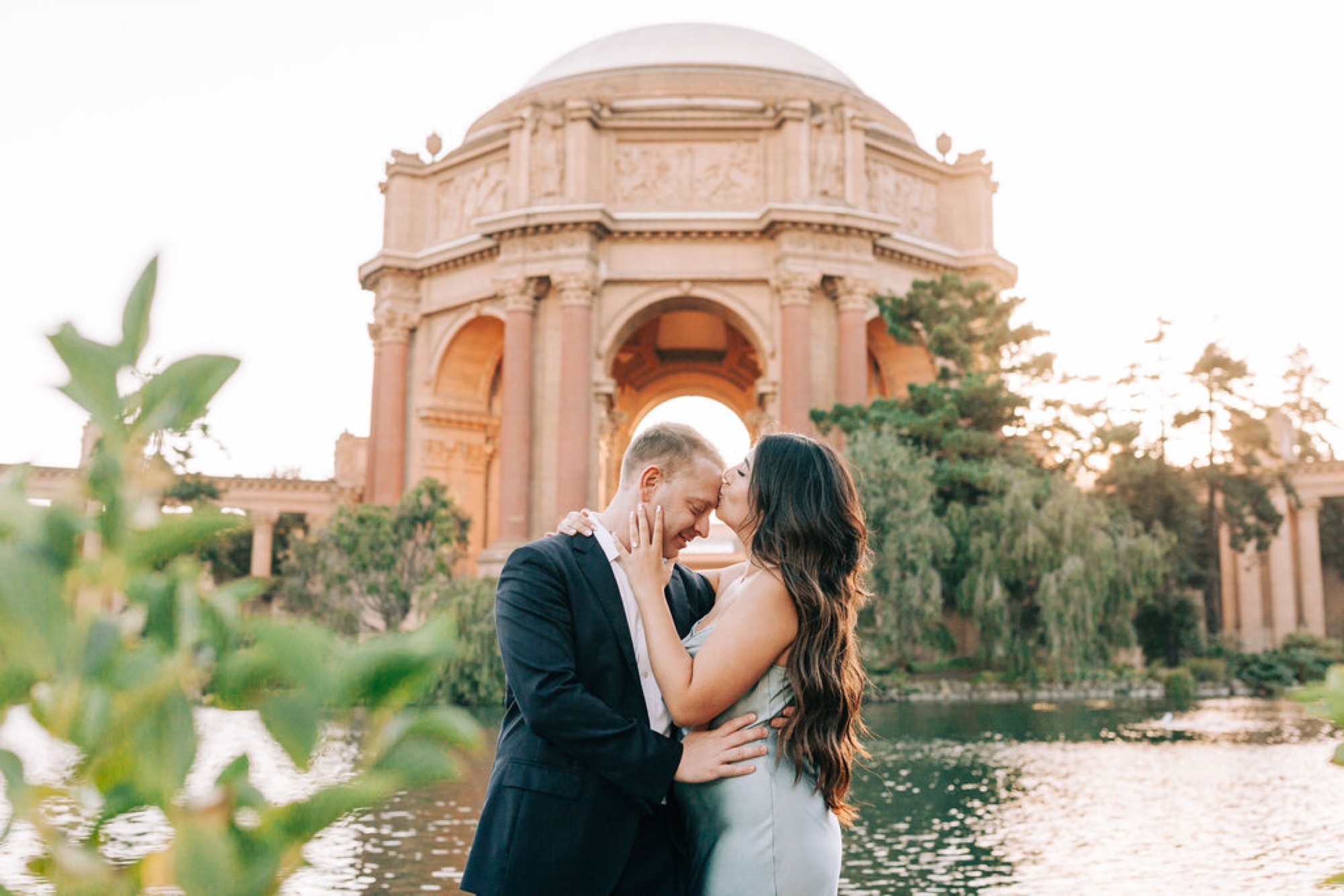 A couple shares an intimate moment at the Palace of Fine Arts, with the iconic dome glowing warmly behind them as golden hour light filters through surrounding greenery