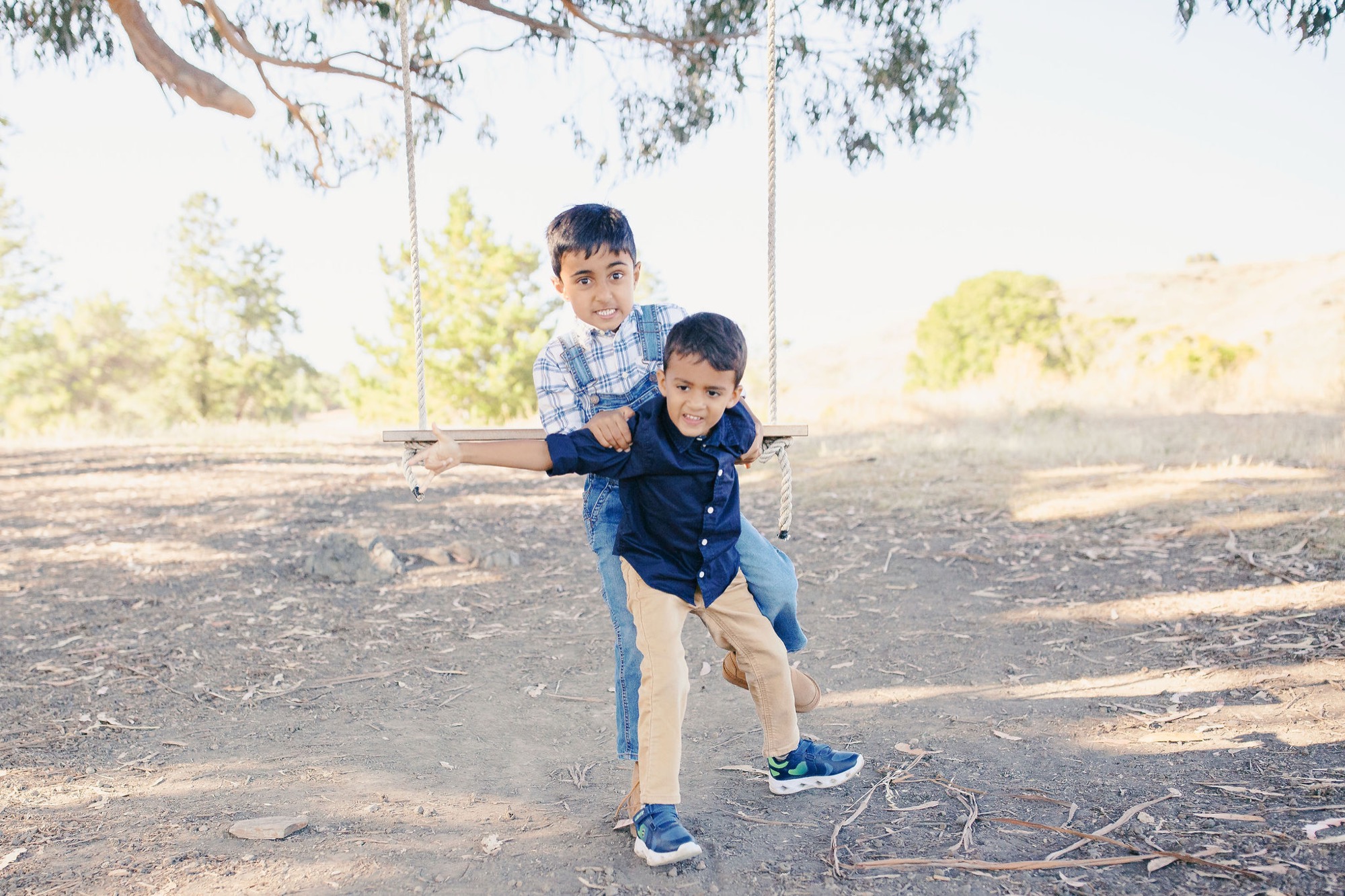 Two young boys play on dusty ground beneath a eucalyptus tree, the older child balanced behind the younger one in concentrated playful pose