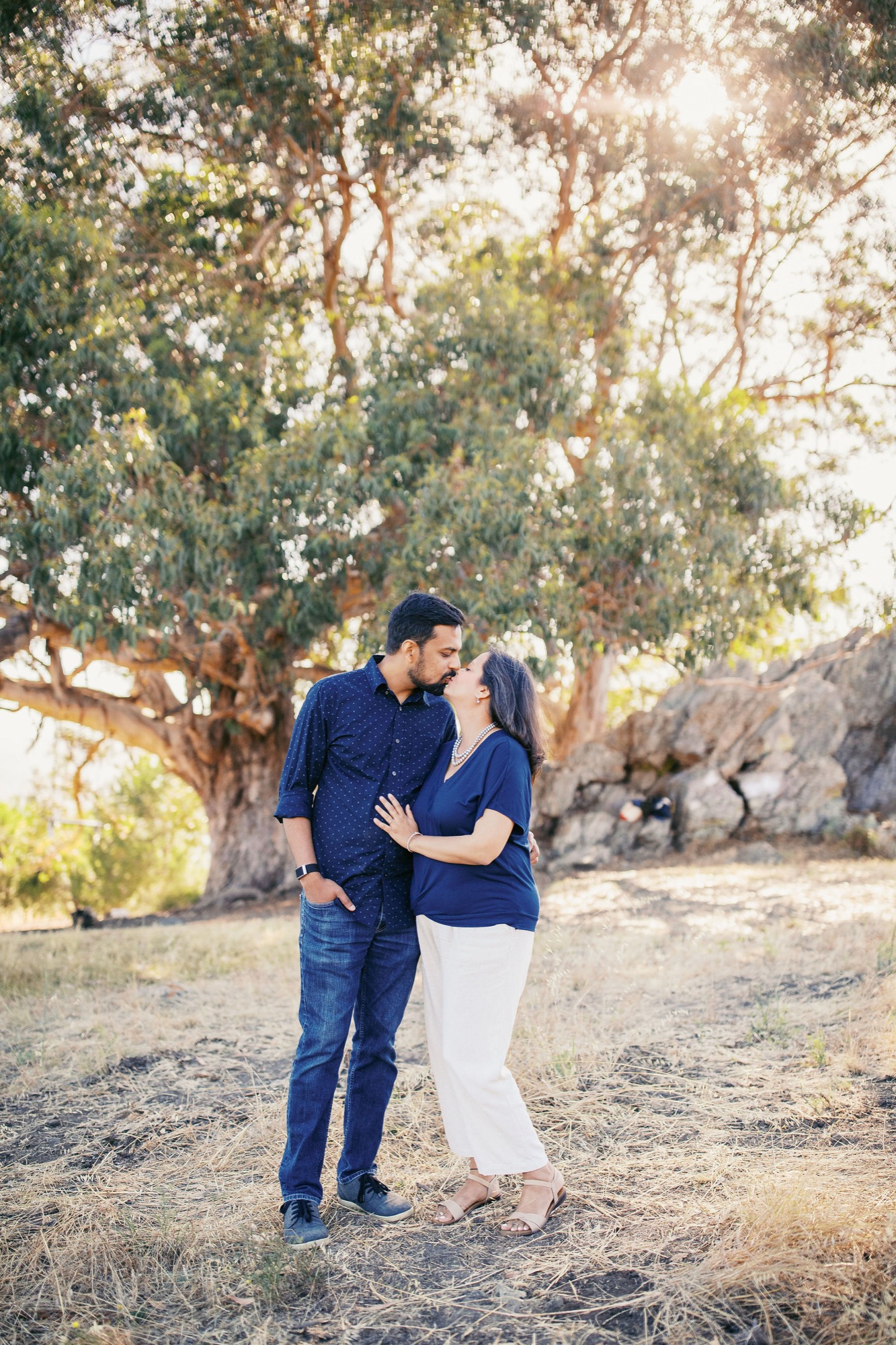 A couple in matching navy tops embraces gently beneath towering eucalyptus trees, golden sunlight filtering through the canopy onto the dusty ground