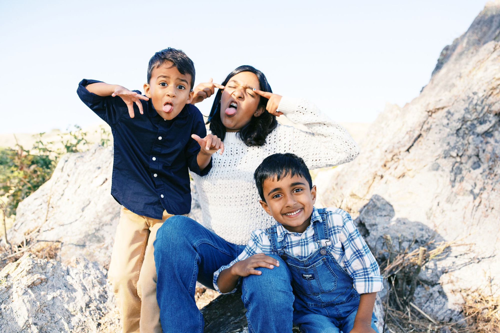 A woman and three boys playfully pose on rocky terrain with windswept hair, bright expressions, and animated gestures against chalky hillside cliffs