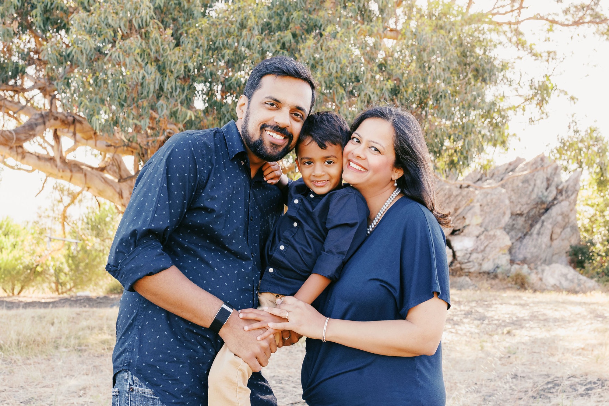 A bearded father, mother, and young son in navy shirts stack hands together under a sprawling oak tree, smiling warmly at the camera with golden afternoon light filtering through the canopy