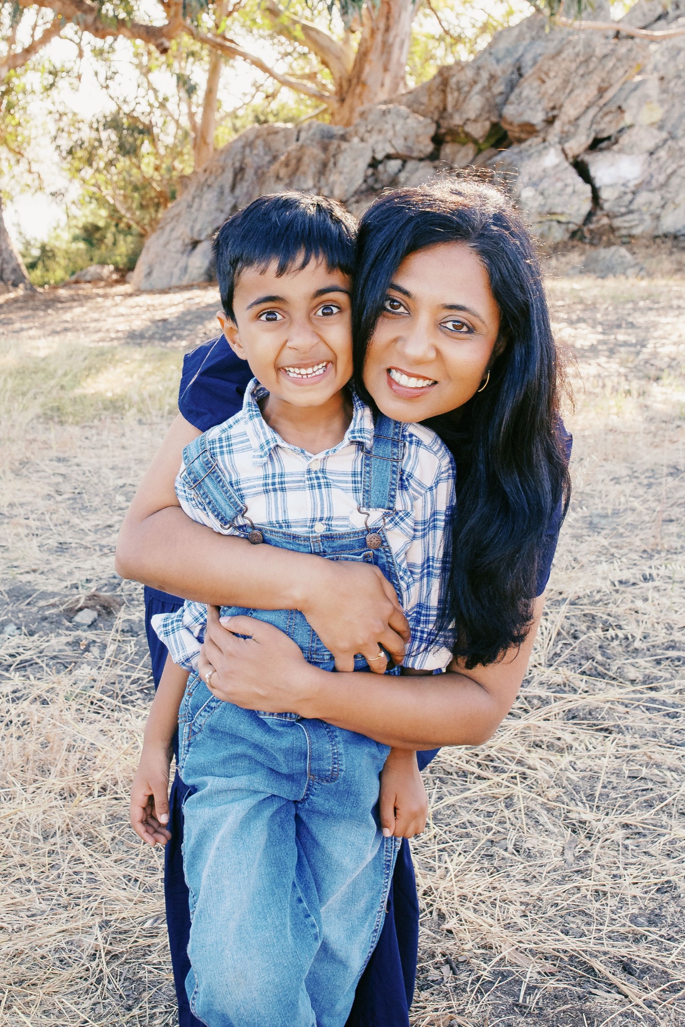 A young woman piggybacks a smiling boy in a blue plaid shirt against a large boulder and eucalyptus trees in dappled sunlight
