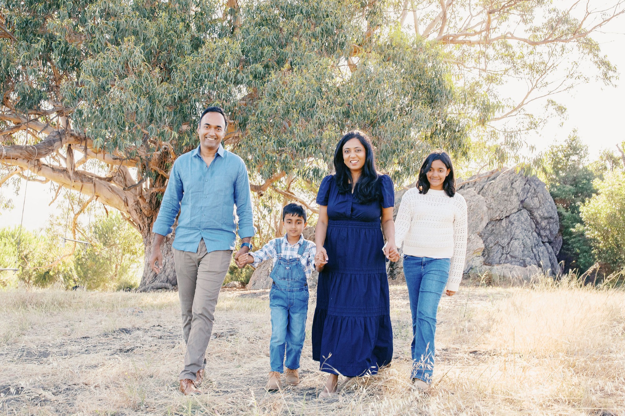 A family of five walks hand-in-hand toward the camera on dusty ground, backlit by golden afternoon sun filtering through sprawling oak trees