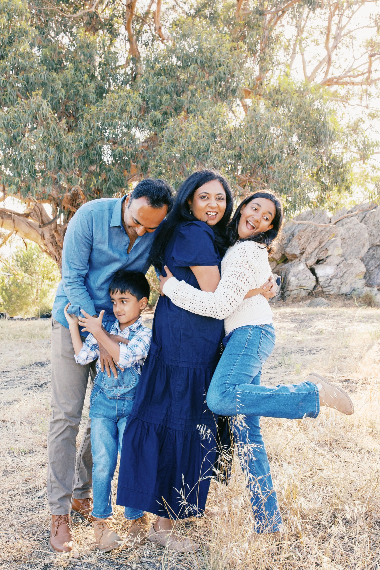 Family of four huddles together laughing against golden hillside trees, wearing casual blues and denim in warm afternoon light