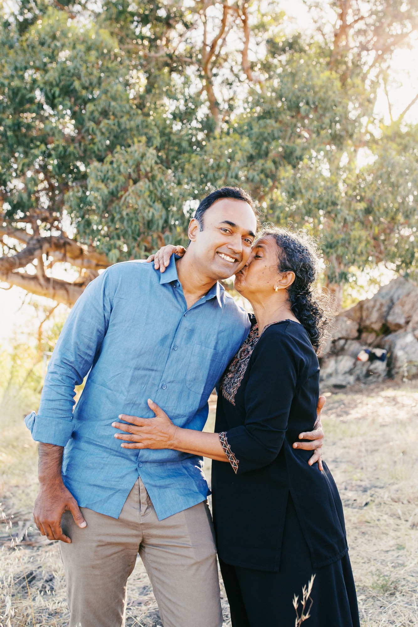 A couple shares a joyful moment among eucalyptus trees, with warm afternoon light filtering through the canopy as she kisses his cheek
