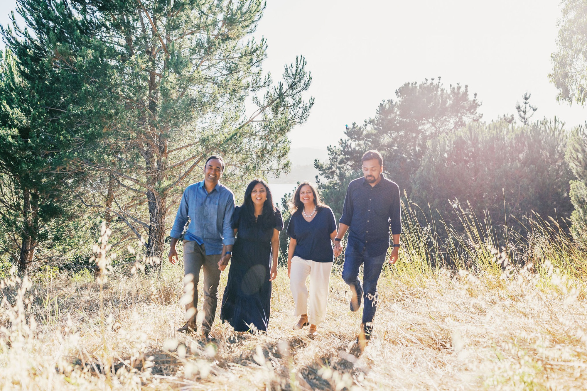 Marin family photographer captures two families walking together through sun-drenched golden grasses and pine trees — laughing, hand in hand, in the soft hazy backlight of a beautiful Marin County afternoon. Extended family lifestyle photography at its most natural and joyful.