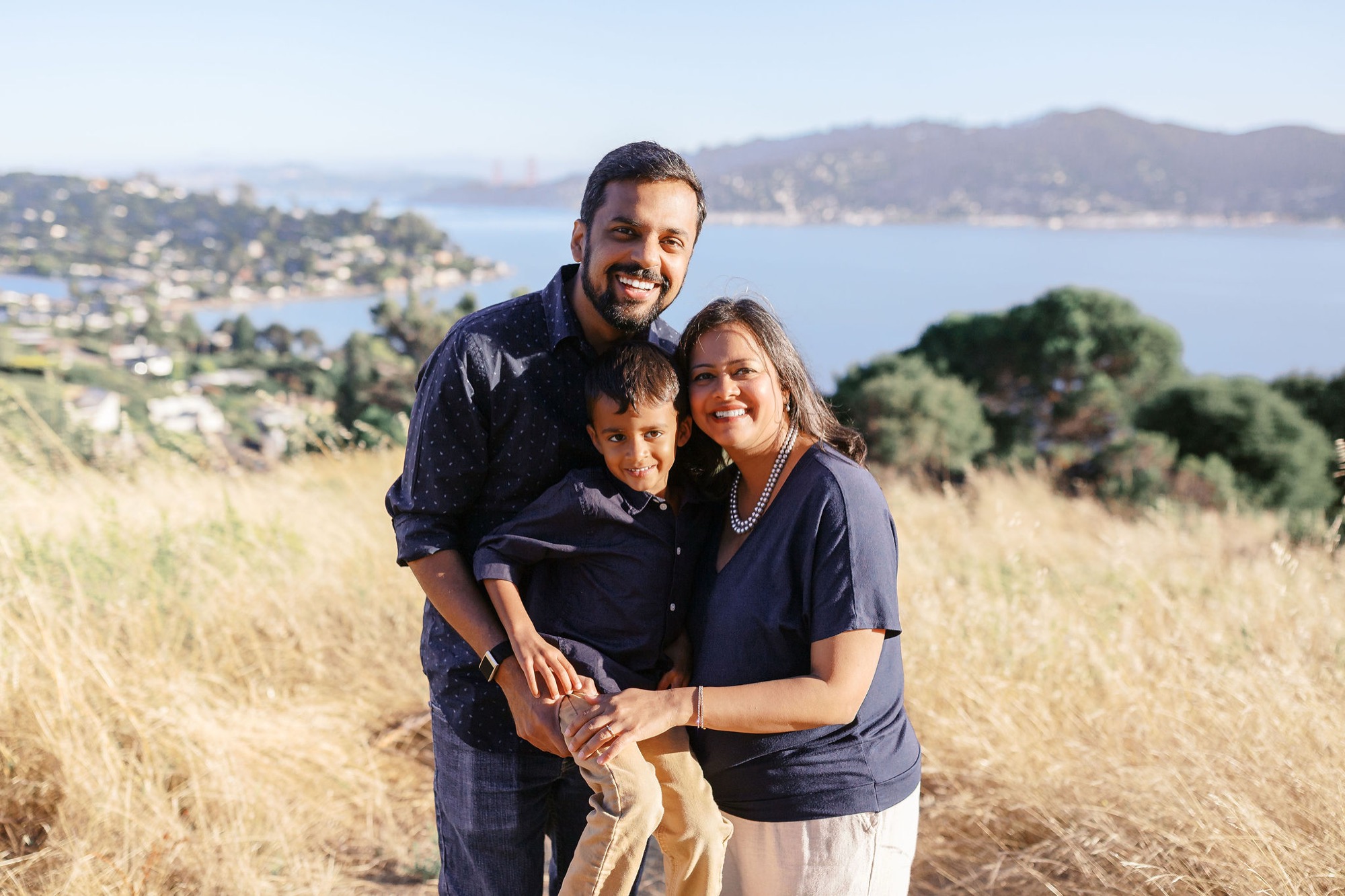 Marin family photographer captures a joyful family of three in coordinated navy against the stunning backdrop of the San Francisco Bay and Golden Gate Bridge — a perfect outdoor portrait session in Marin County's golden hills.