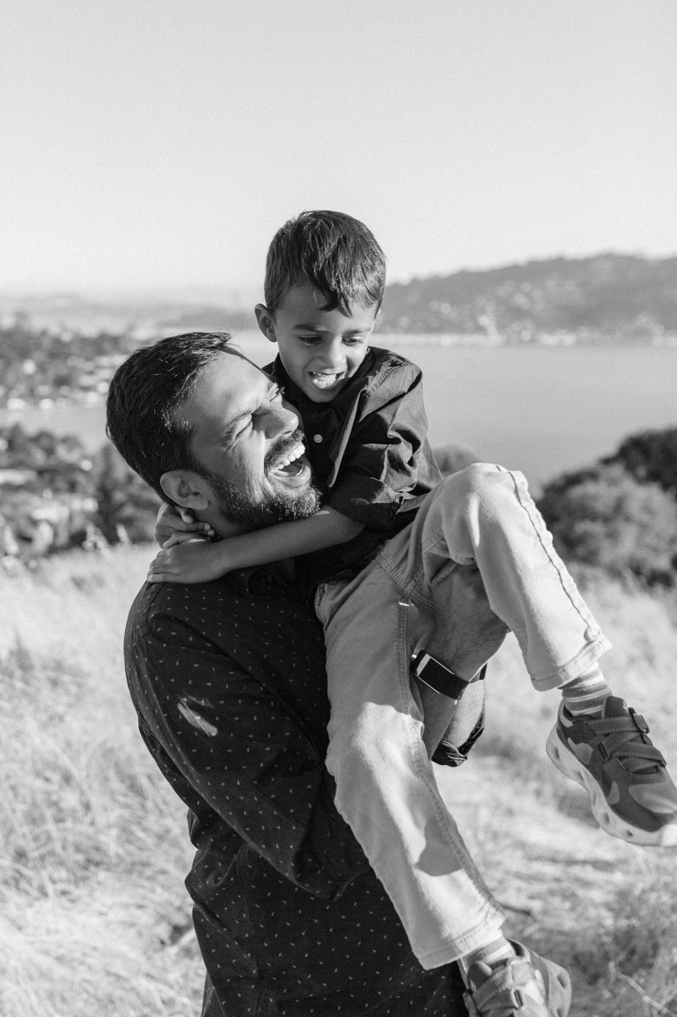 Marin family photographer captures a father erupting in laughter as he lifts his joyful son on a sun-drenched hillside overlooking the Marin County bay — authentic candid family photography at its best.