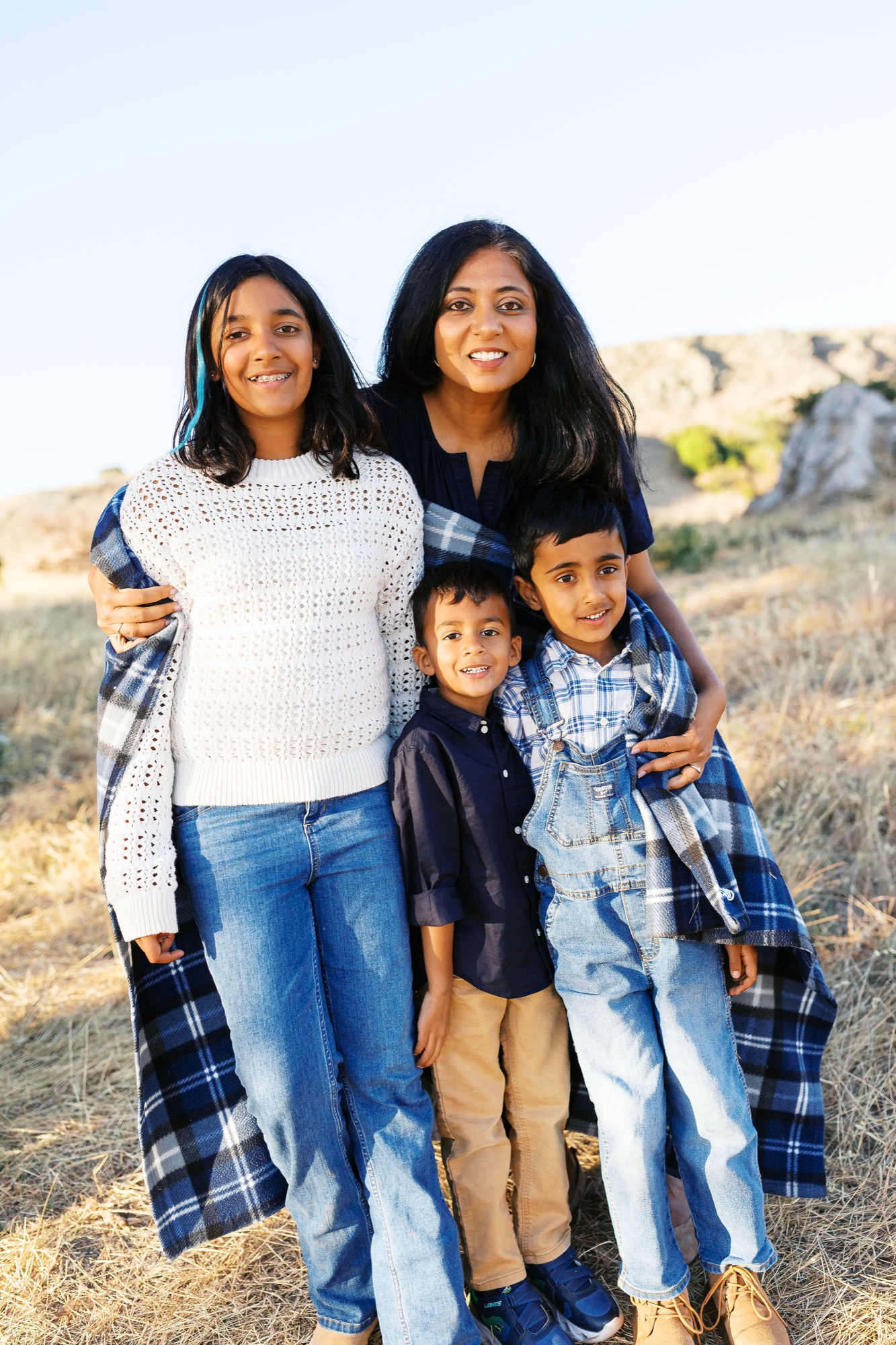 Marin family photographer captures a warm and radiant family portrait in the golden outdoor light of Marin County — mom and her three children huddle close together, smiling naturally against a backdrop of dry hills and clear blue sky in this beautiful Bay Area family session.